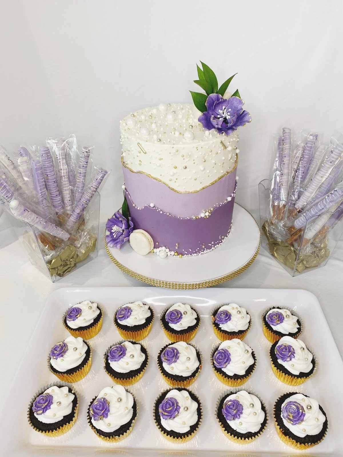 A cake with purple and white icing decorated with gold accents and purple flowers, surrounded by cupcakes with white icing and purple flowers, and two baskets of rolled cookies in clear plastic packaging.
