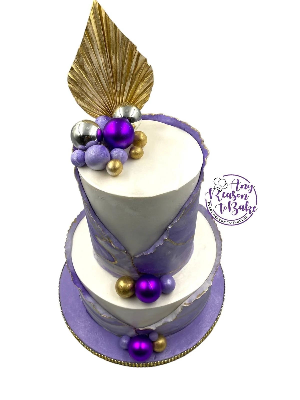 A two-tiered cake decorated in purple, white, gold, and silver with a gold leaf and purple ornament topper, and topped with metallic purple, silver, and gold spheres. The cake has a purple base with gold accents and a gold circle border on the bottom tier. There is a logo with a chef's hat that reads 'Any Reason to Bake' on the side.