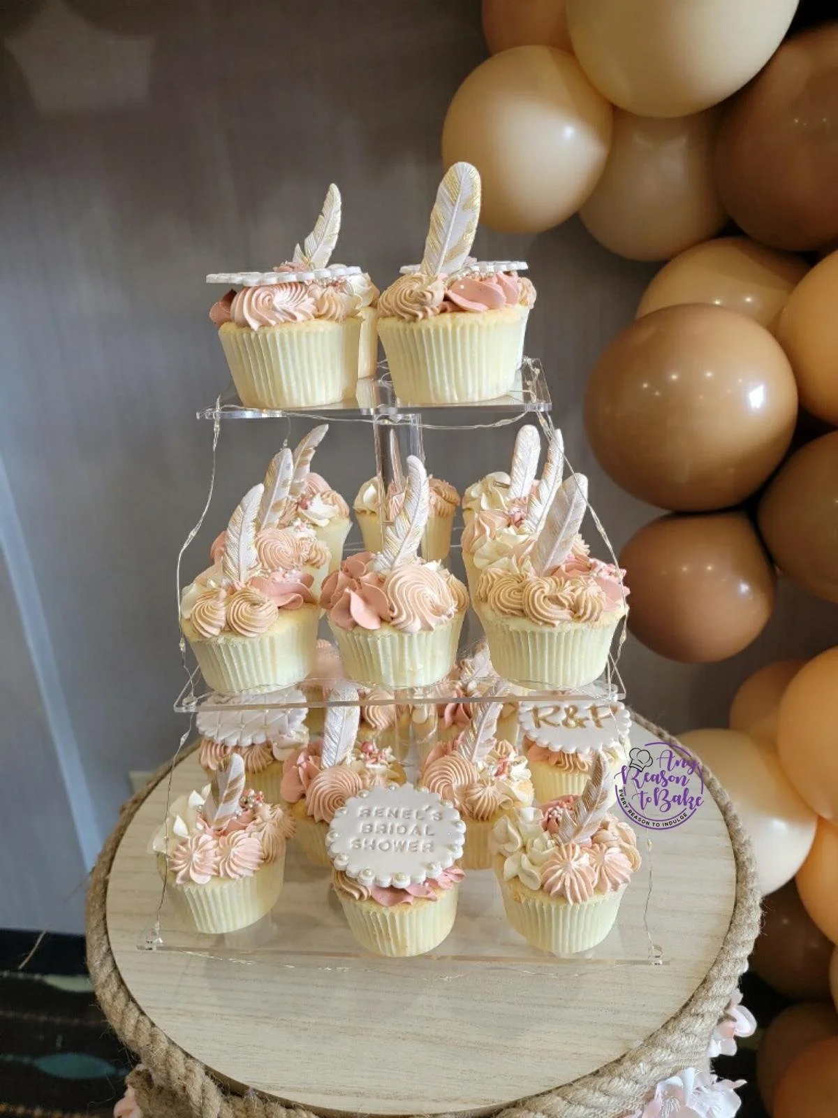 Cupcakes decorated with light pink and white frosting, feather toppers, and a small sign with the message, 'René's Bridal Shower'. The cupcakes are arranged on a three-tiered clear stand on a rustic wooden table with beige and brown balloons in the background.