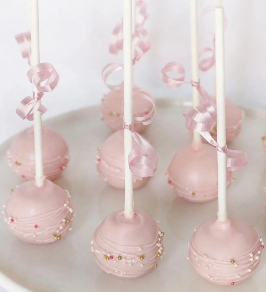 Pink cake pops decorated with small colorful sprinkles and tied with pink curly ribbons, arranged on a white stand.