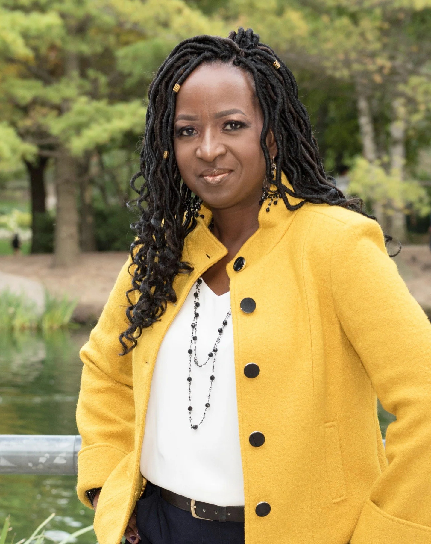 A woman with long black dreadlocks wearing a bright yellow coat with black buttons, a white blouse, and layered black necklaces standing outdoors near a pond with green trees in the background.