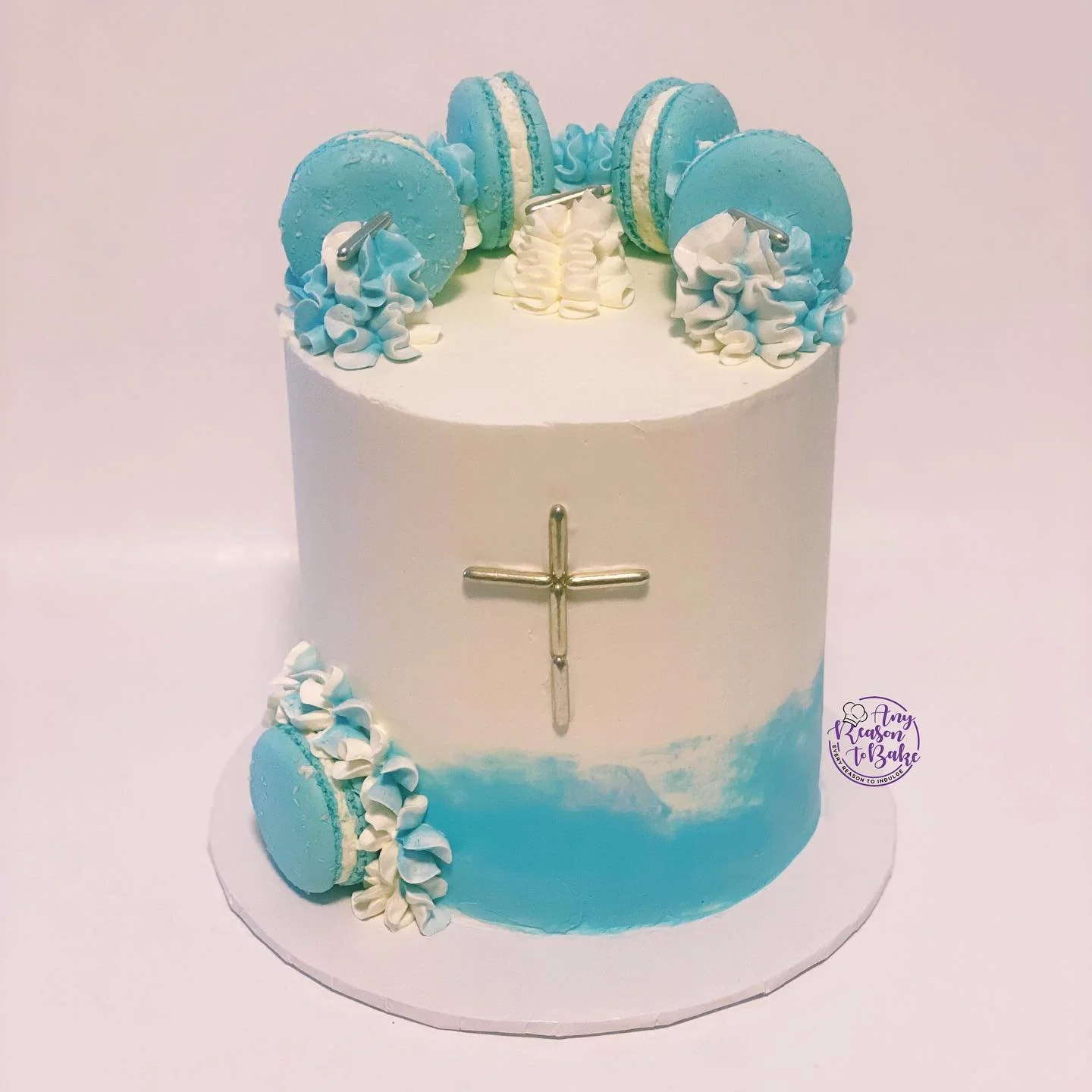 A two-tiered cake decorated with blue and white frosting, featuring blue macarons on top and around the base, and a silver cross on the front, with a small purple logo of "Amy Reason to Bake" on the side.