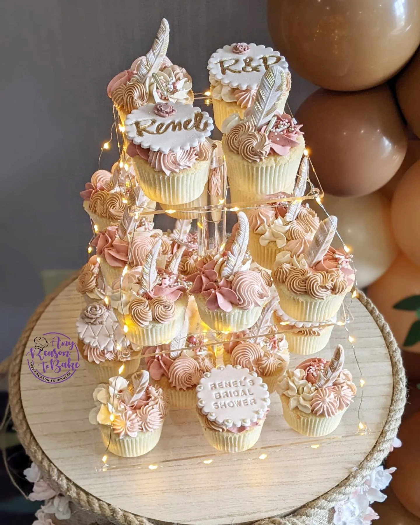 A tiered cupcake stand displaying pink and white decorated cupcakes with feathers, pearls, and floral accents. Some cupcakes have writing such as "Remily" and "K&R" on white fondant decorations, indicating a bridal shower celebration.