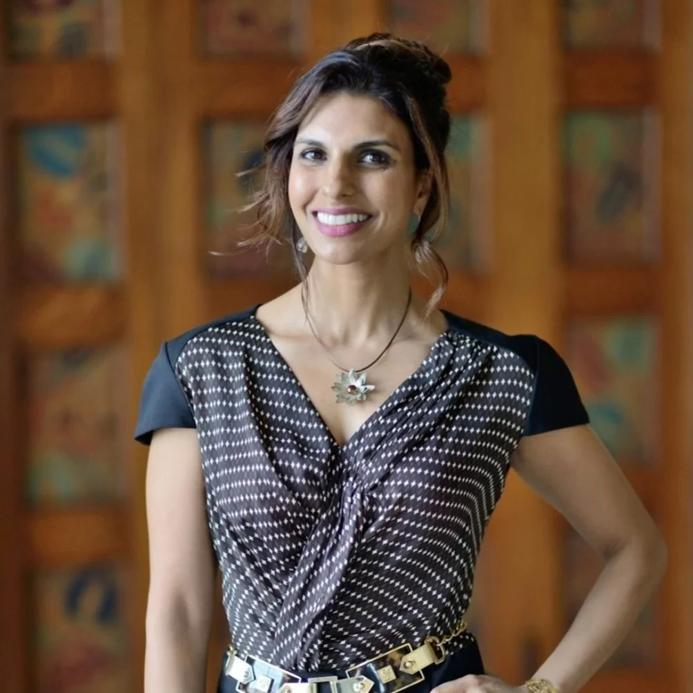 A woman with shoulder-length brown hair, wearing a black and white polka dot dress, smiling, standing in front of a wooden background.