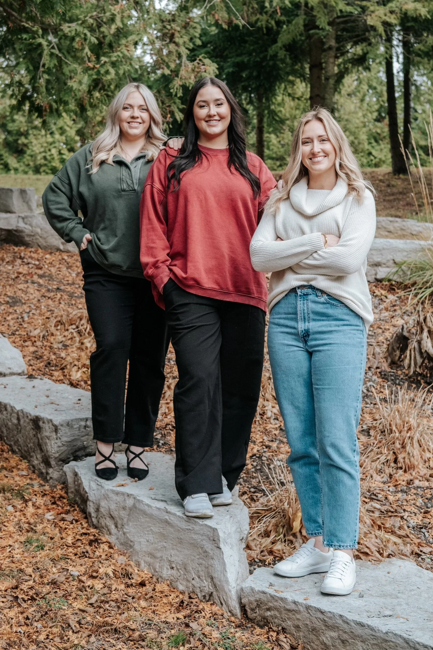 Three women standing outdoors on a stone pathway surrounded by fall foliage and trees, smiling at the camera.