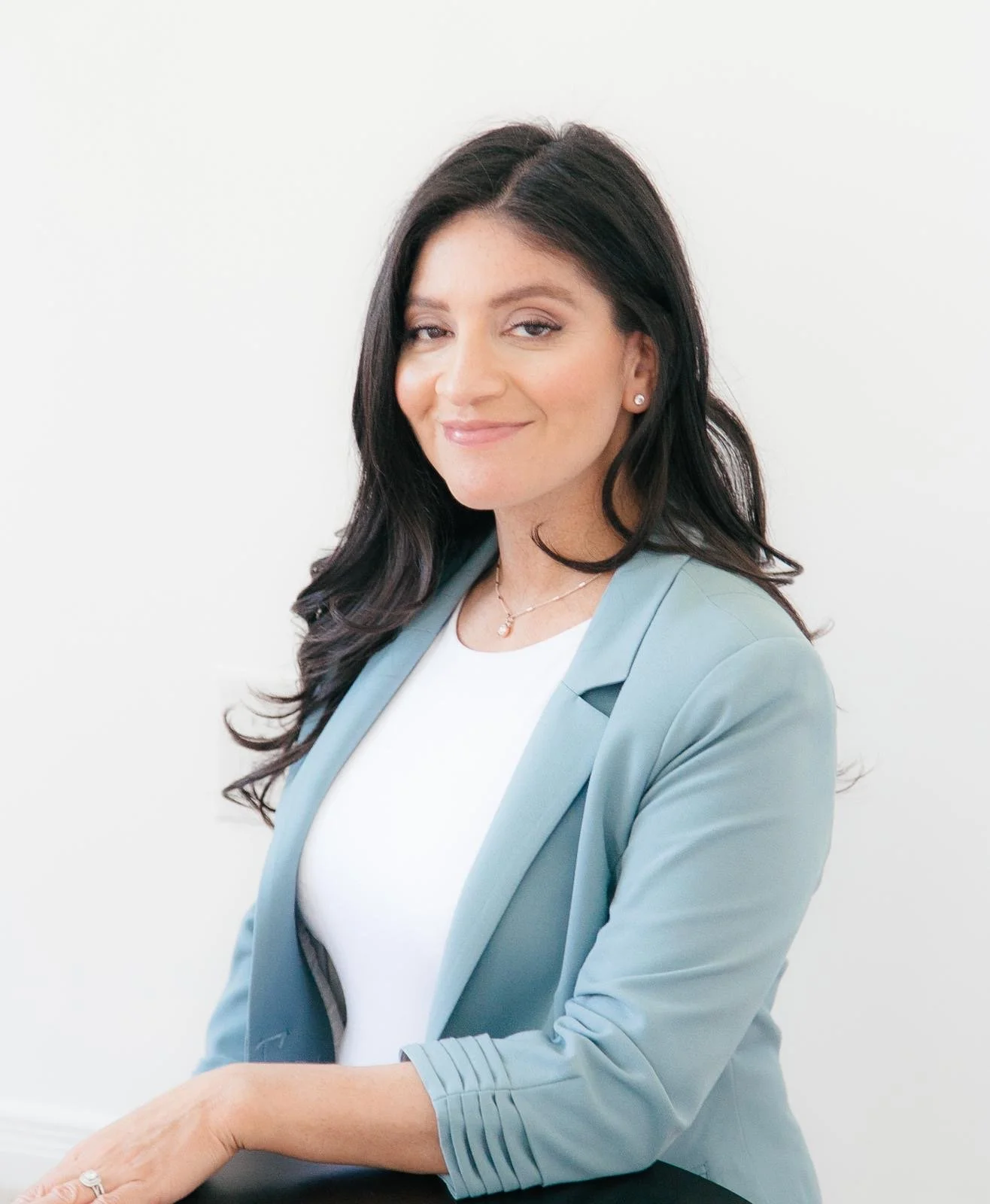 Headshot of a woman with long dark hair wearing a light blue blazer and white top, smiling and standing against a plain white background.
