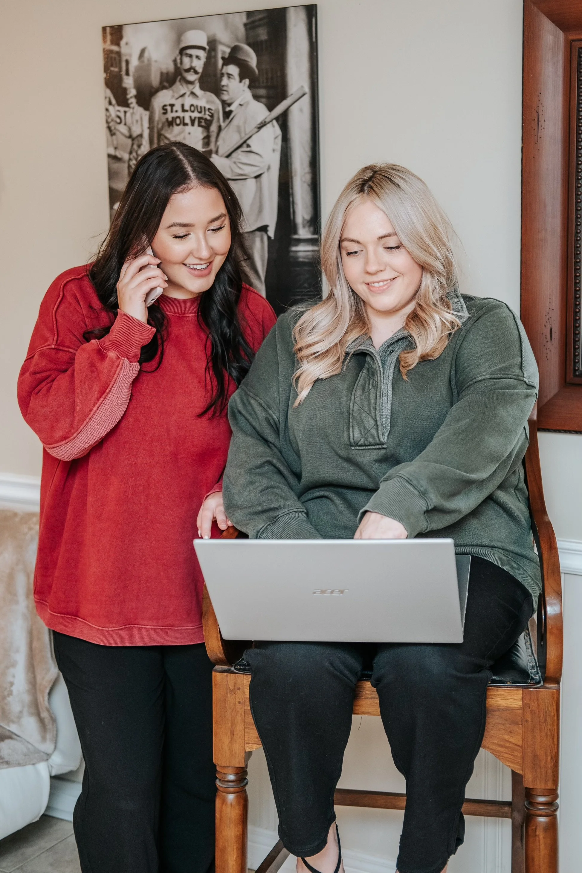 Two women, one sitting on a wooden chair and the other standing close, looking at a laptop screen with smiles. The woman sitting is blonde with a green oversized sweatshirt, and the standing woman has dark hair, wearing a red long-sleeve shirt. Behind them is a black and white vintage photo on the wall.