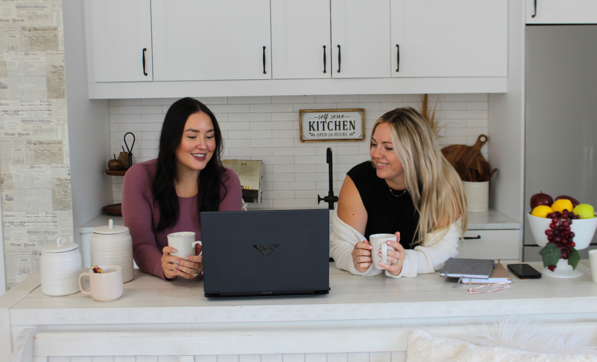 Two women sitting at a kitchen counter, smiling, with a laptop open in front of them and holding mugs. The kitchen has white cabinets, a fruit bowl with apples and grapes, and a sign that reads 'self serve KITCHEN OPEN 24 HOURS' in the background.
