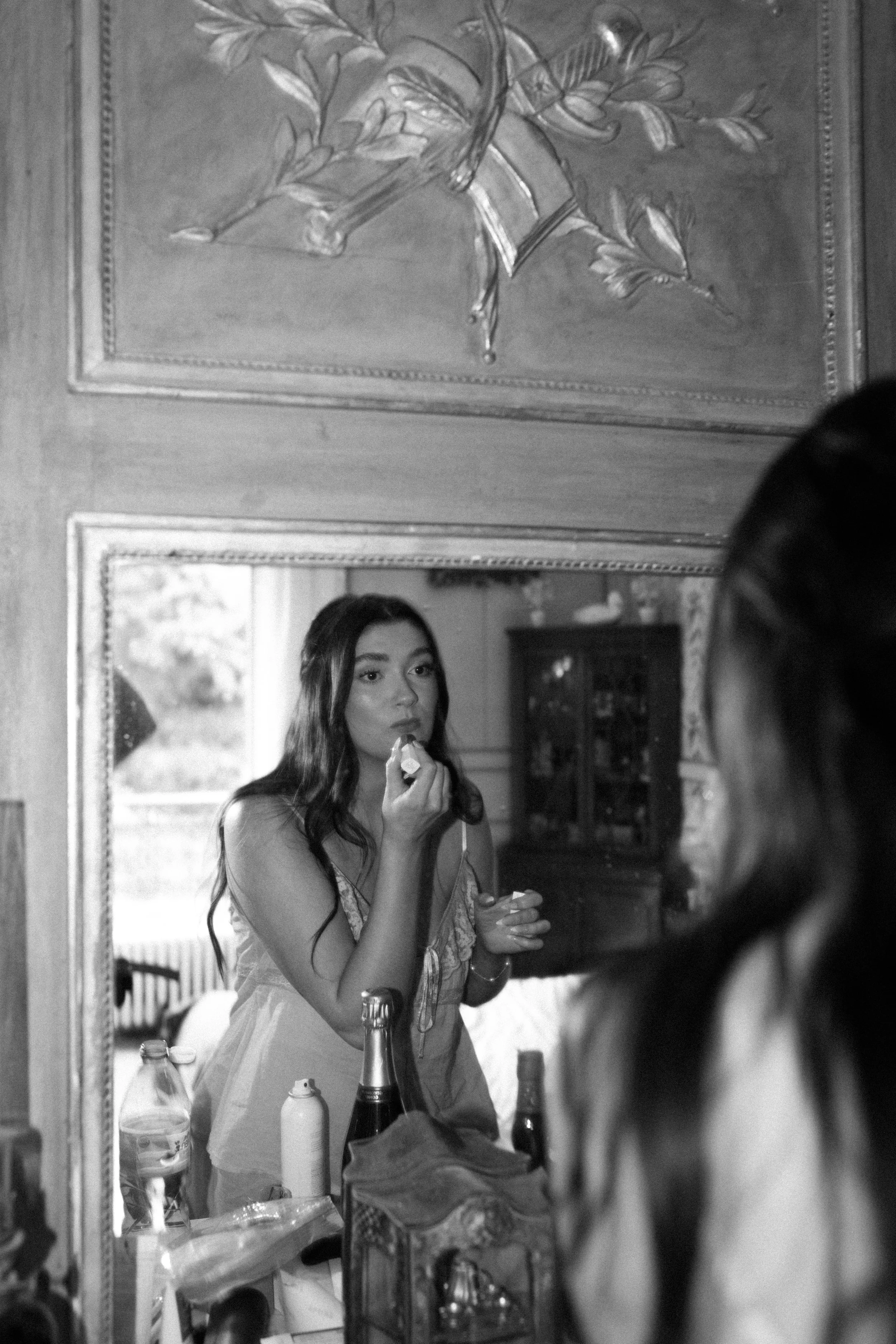 Black and white image of bride getting ready, applying lipstick in front of a mirror with soft natural light highlighting an intimate and elegant wedding morning moment