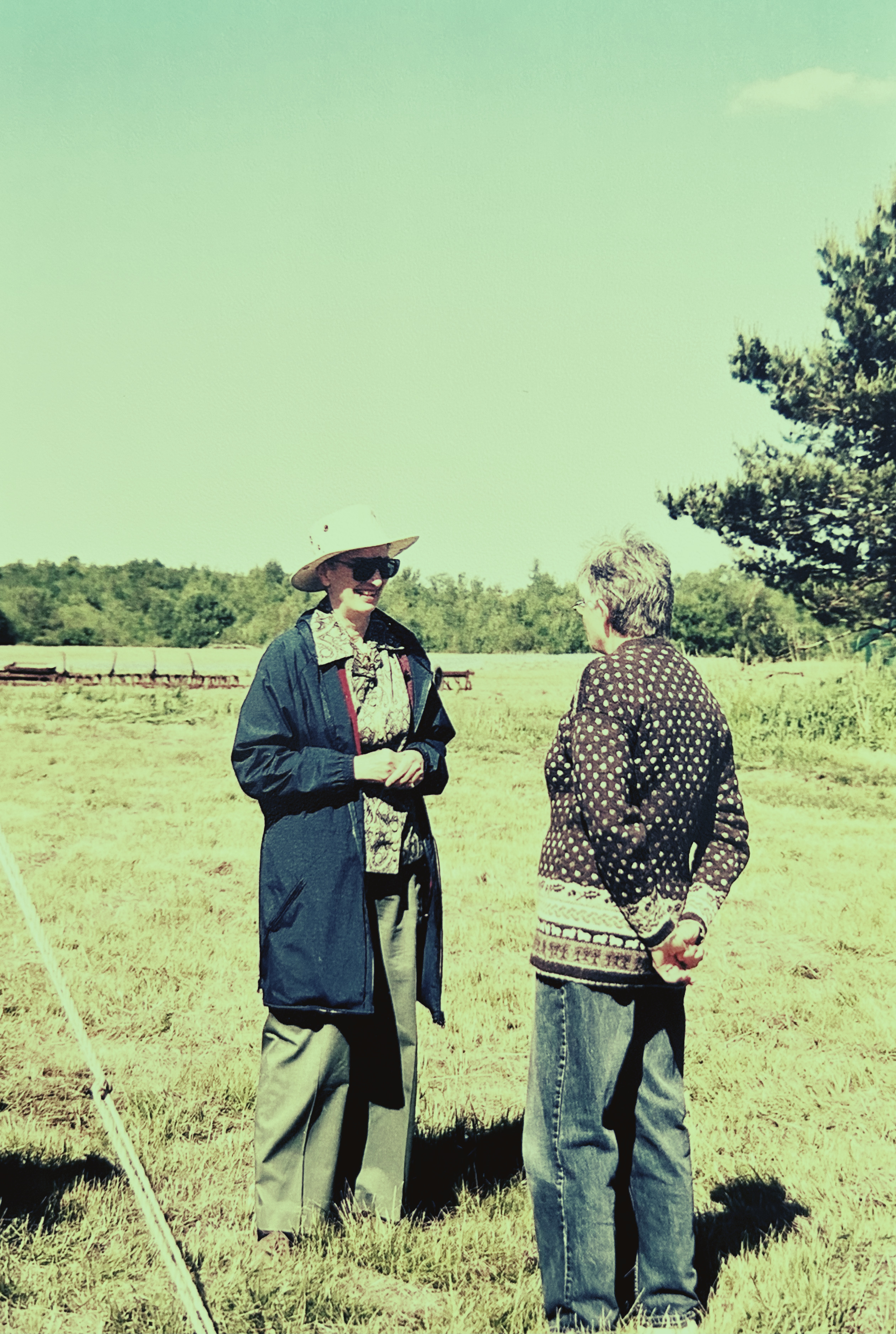 Two older women standing in a field facing each other, one with hands behind her back and one clasped in front of her.
