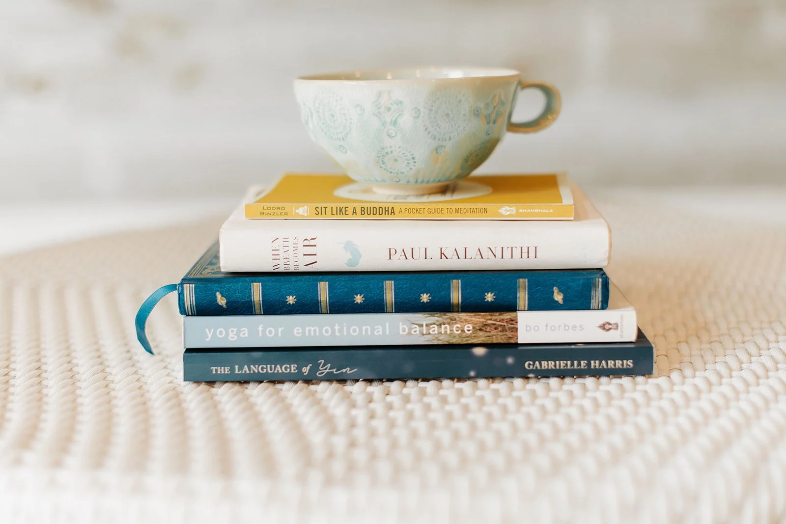 Stack of five books with a ceramic cup on top, placed on a textured surface. The books include titles about meditation, emotional balance, language, and yoga.