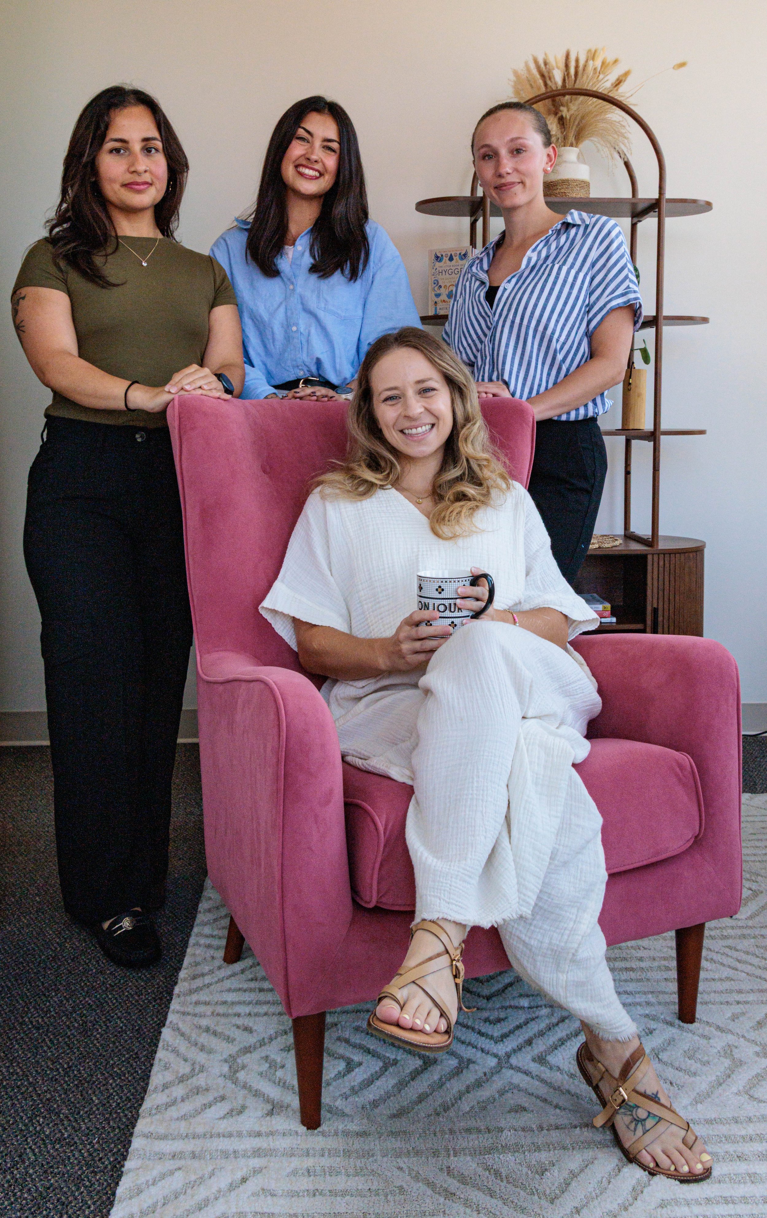 Four women in a living room, one sitting in a pink armchair holding a cup, three standing behind it, with a bookshelf and decorative plants in the background.