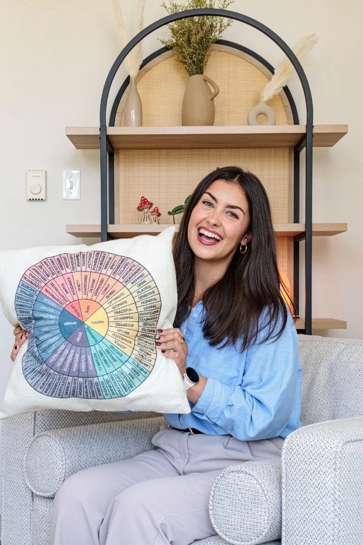 A woman sitting on a white sofa holding a colorful wheel chart pillow, smiling at the camera in a well-lit living room with decorative vases and art on shelves behind her.