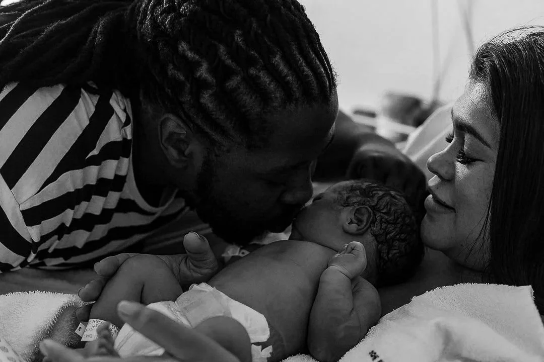 A man and woman holding a newborn baby in a hospital bed, with the man kissing the baby's forehead and the woman looking lovingly at the baby.