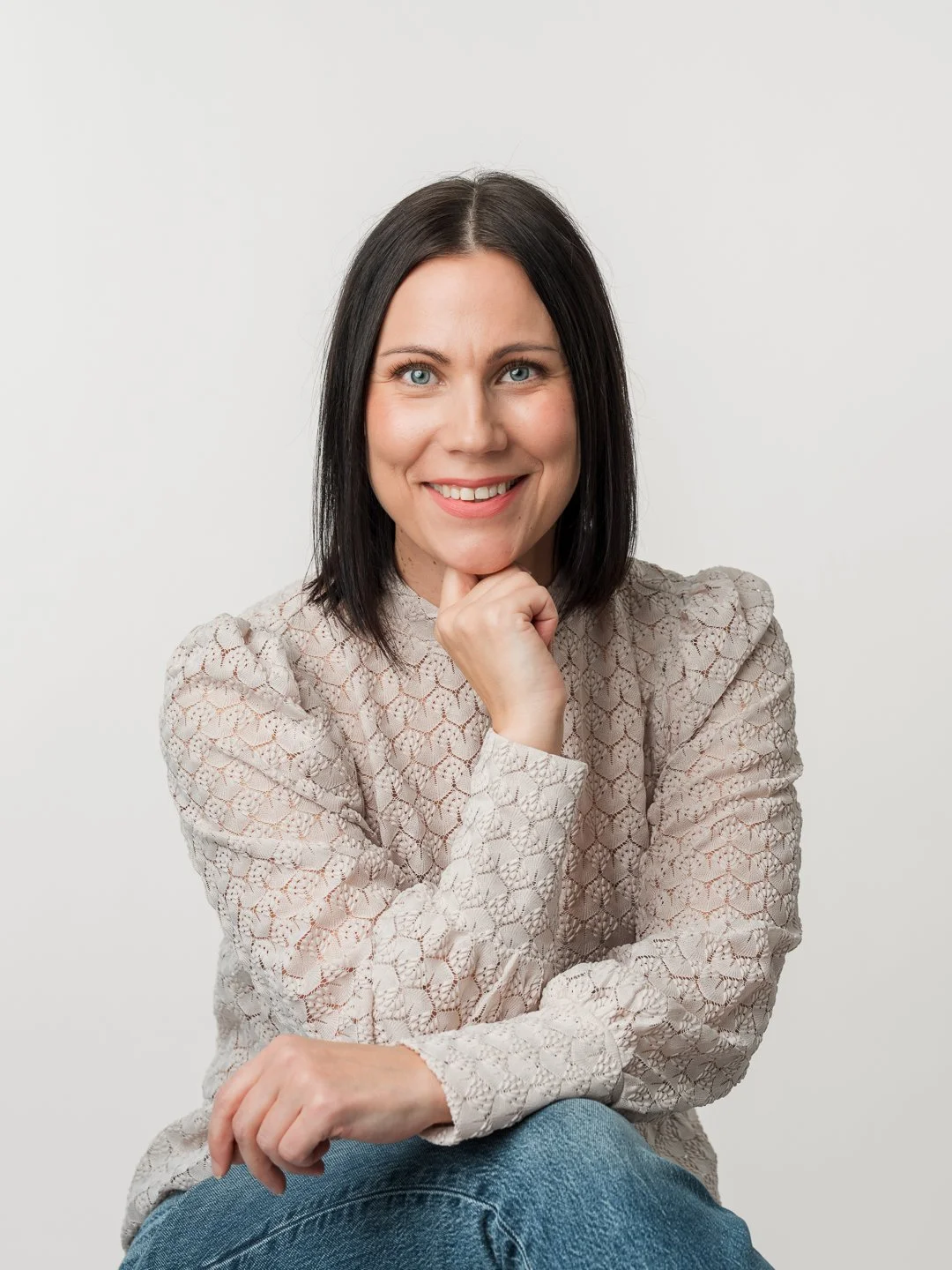 Harmaavalkoinen sorrava nainen, jolla on vaaleat silmät, piilottaa suunsa leveään hymyyn, istuu valkoisella taustalla ja katsoo kameraan.