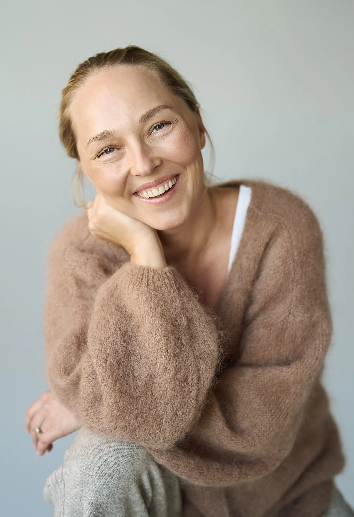 Hymyilevä nainen, istuu nojaten poskeensa kädellä, beige takki ja harmaat housut, vaalea tausta.