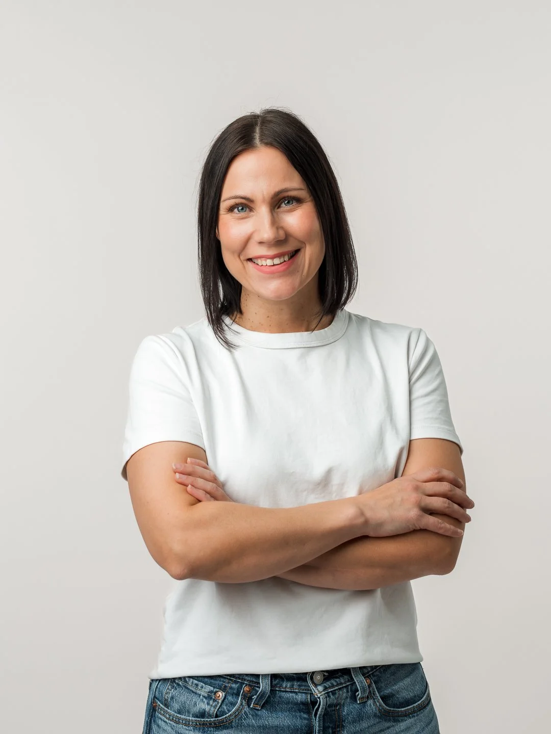 Henkilö, noin 30-vuotias nainen, hymyilee, lyhyt tummatukka, valkoinen T-paita ja farkut, seisoo valkoisella taustalla.