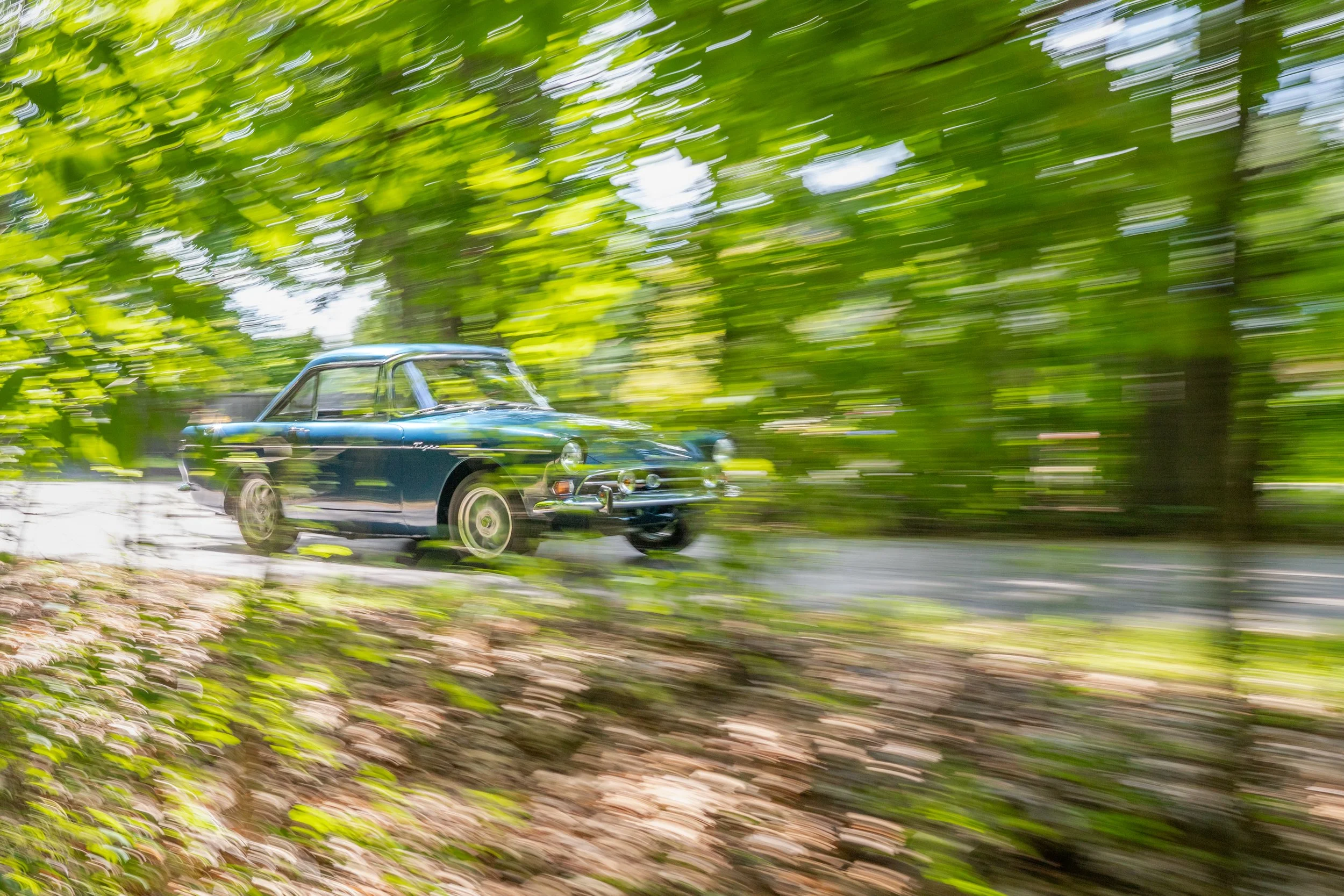 A classic blue car driving through a lush green forest, with motion blur indicating speed.