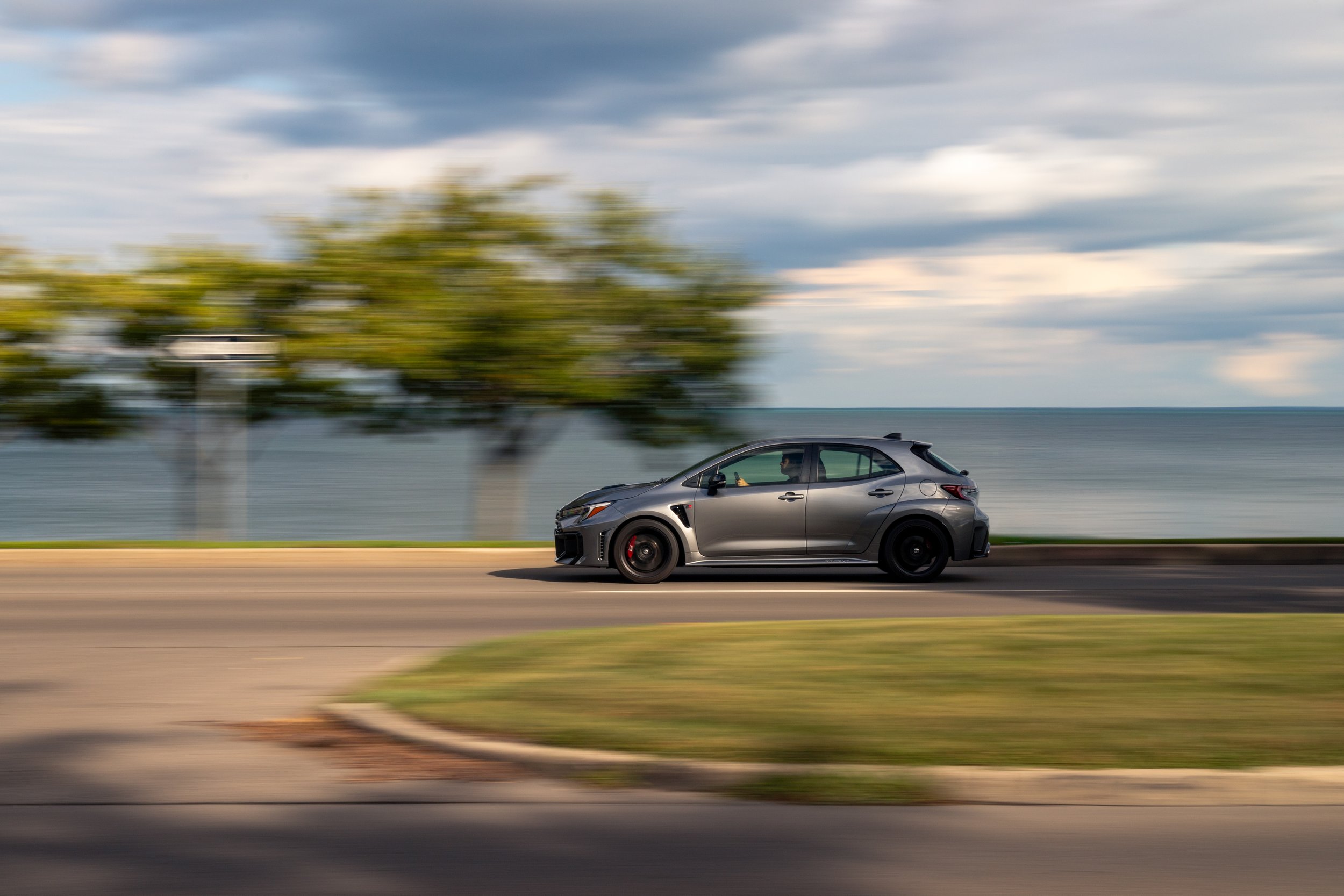 A gray sports car driving along a road by the water with trees and a cloudy sky in the background.