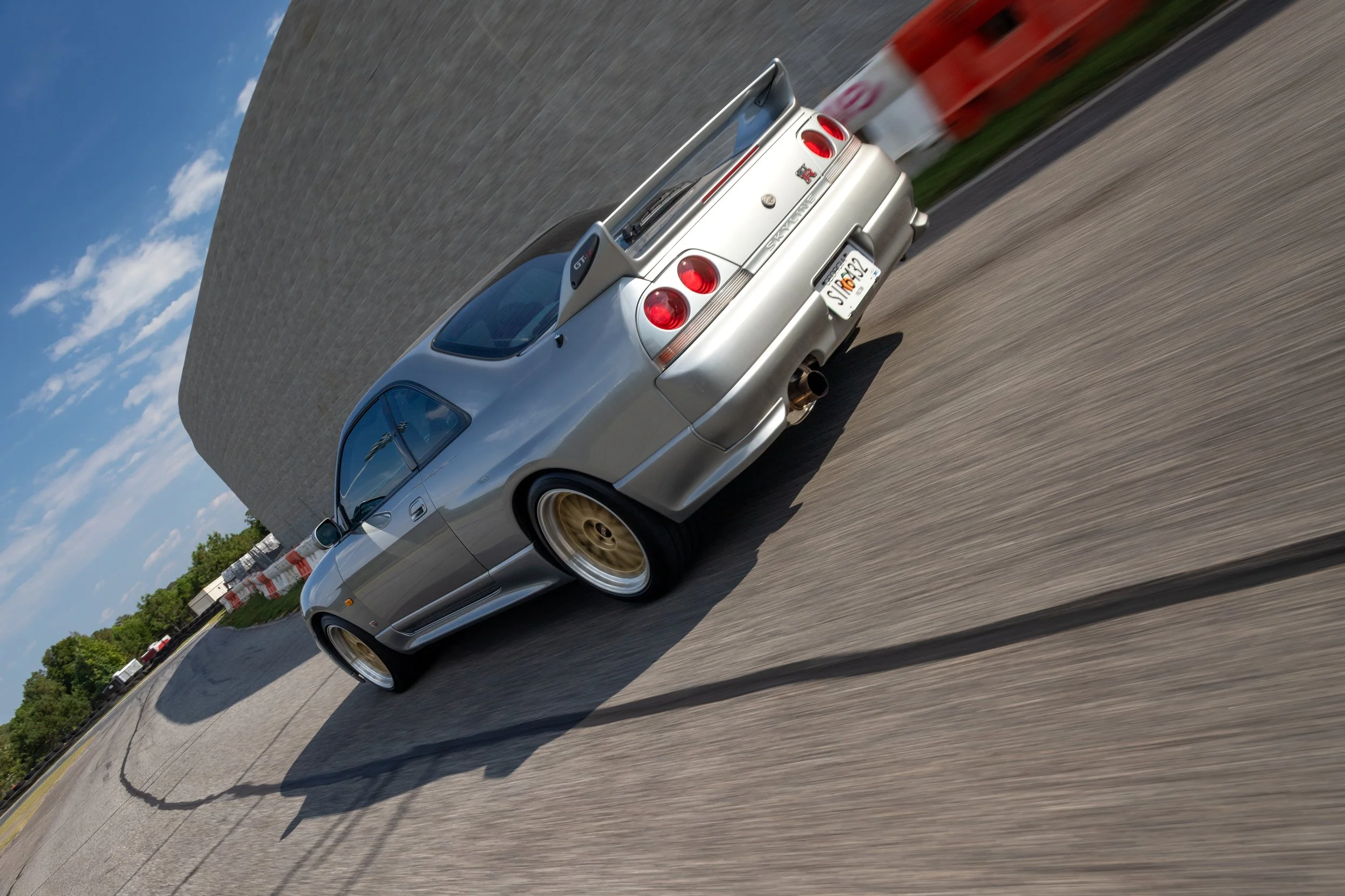 Silver sports car with gold wheels and a rear spoiler driving on a highway near a large concrete wall under a blue sky with some clouds.