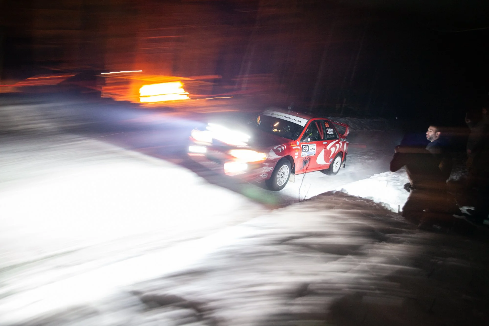 A red rally car with the number 98 on the side, driving at night on snow-covered terrain, illuminated by headlights with a background of sunset or sunrise sky and some spectators watching.