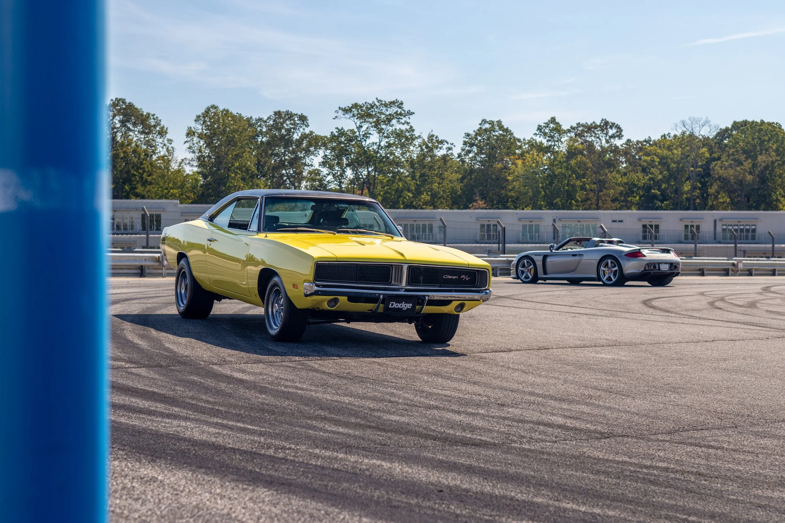 A yellow Dodge Charger R/T parked on an asphalt surface near a chain-link fence, with a silver sports car in the background and green trees under a clear sky.