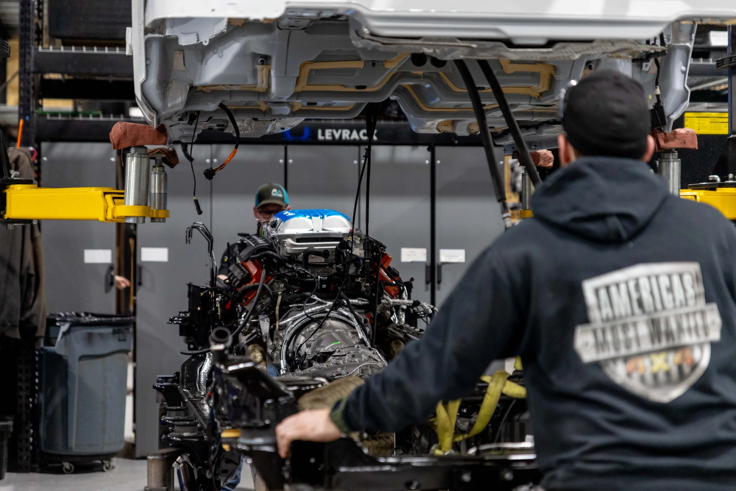 Two workers in a factory or workshop assembling or repairing a complex machine or vehicle component. The workspace includes industrial shelving, yellow lifting equipment, and tools.