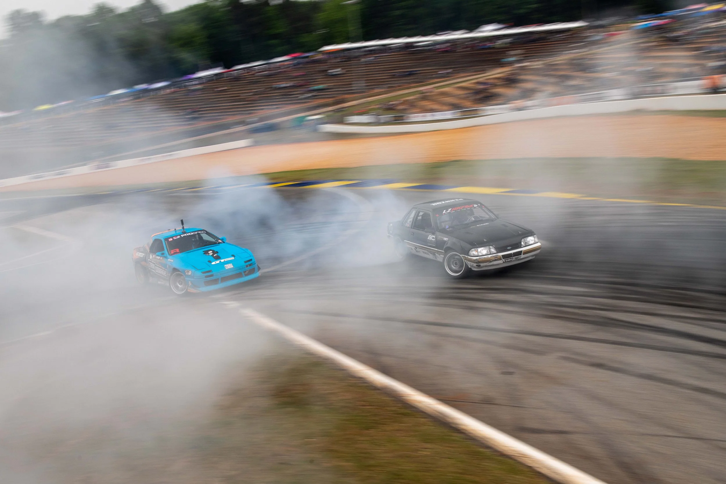 Two cars drifting and smoking tires on a racetrack during a motorsport race, with a blurred audience in the background.