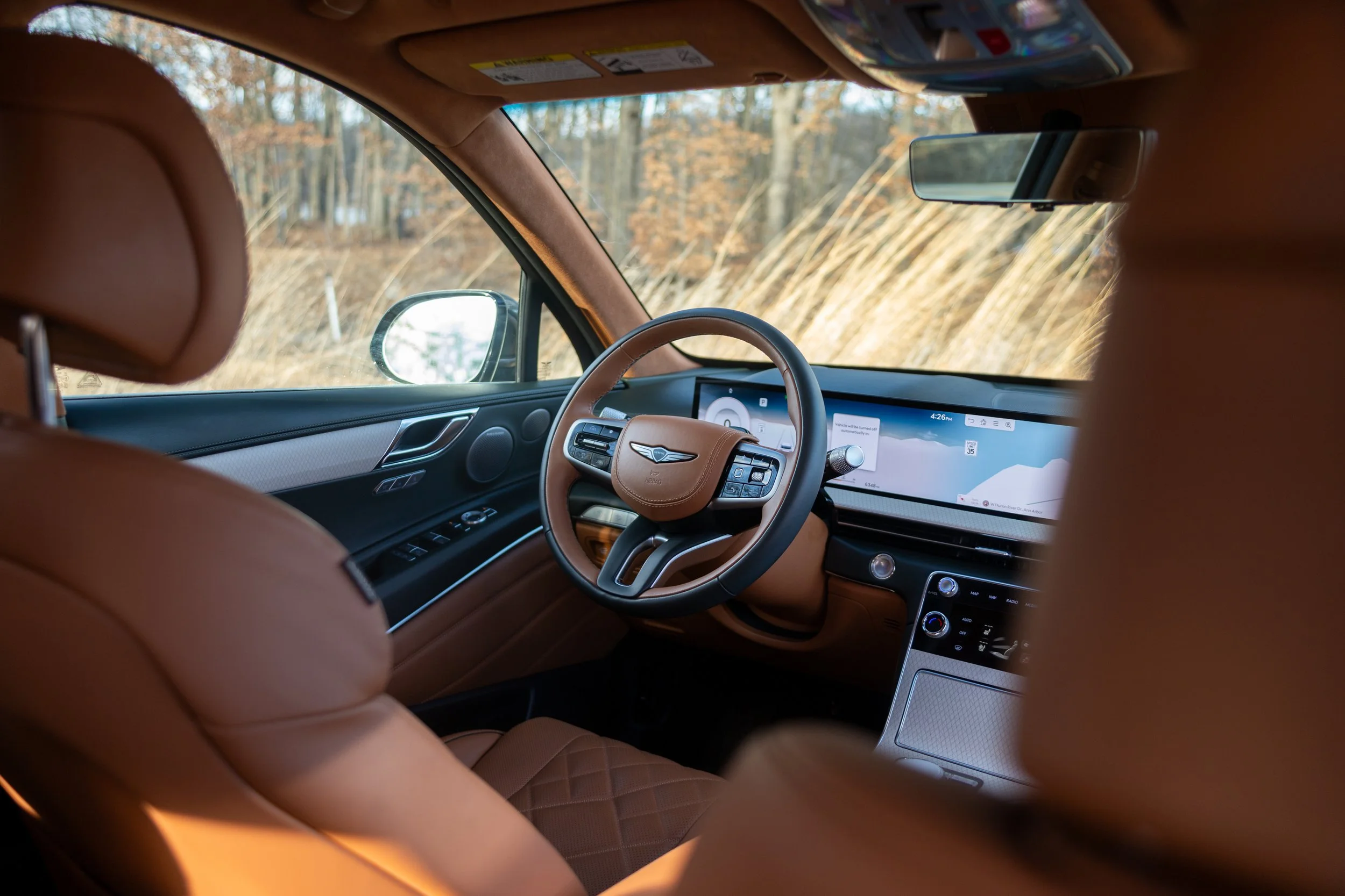 The interior of a luxury car with brown leather seats, steering wheel, dashboard, and a digital display screen, with a view of trees outside the window.