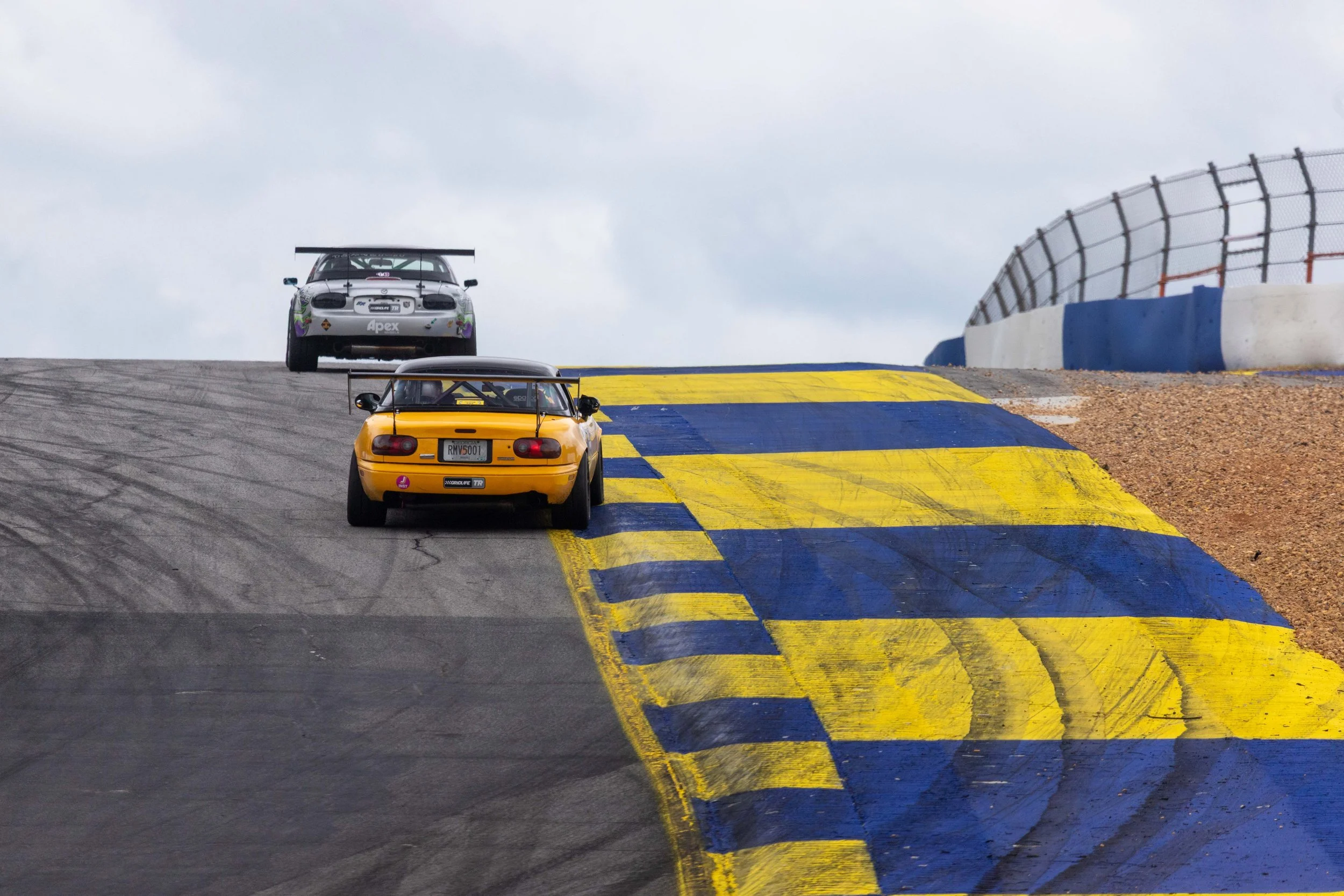 Two race cars on a racing track with a yellow and blue painted section, metal fencing, and a cloudy sky overhead.