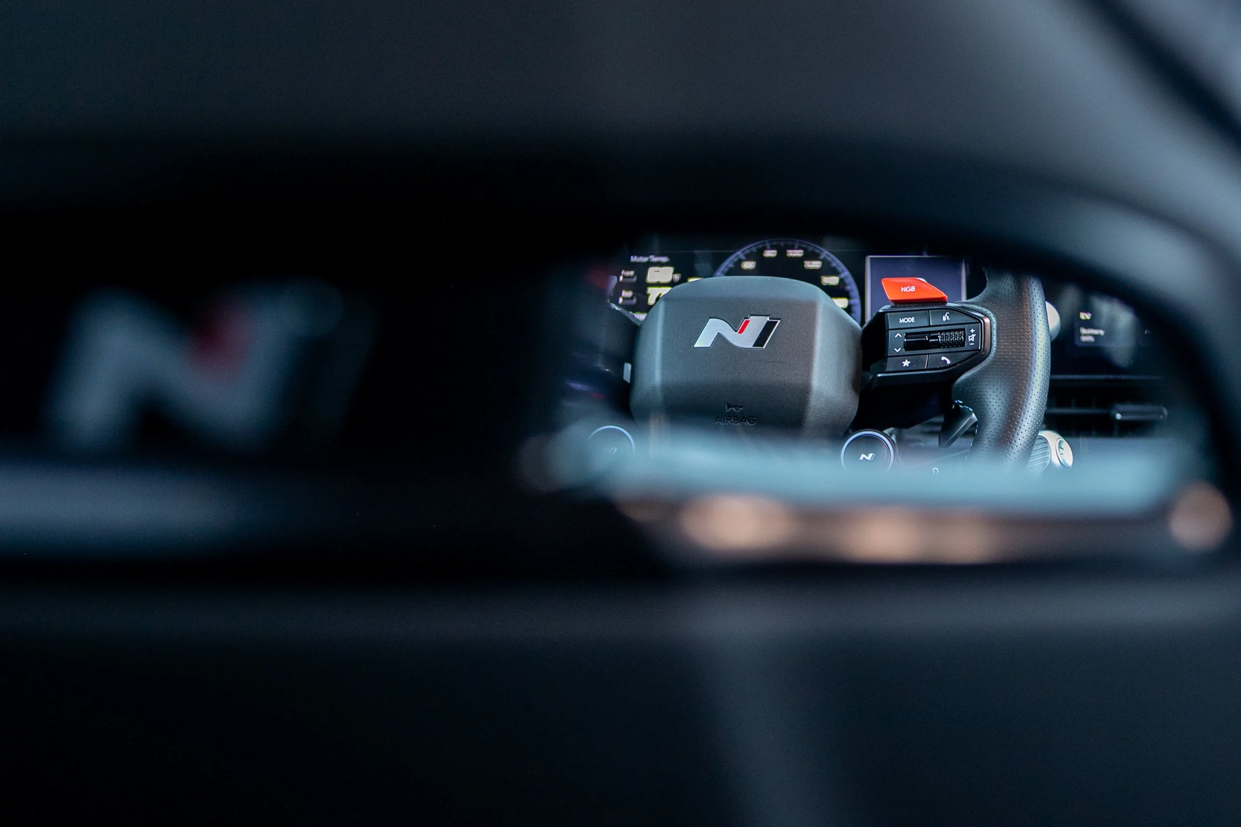 View of a car dashboard with a steering wheel featuring an 'N' logo, digital instrument cluster in the background, and a red 'NGB' button on the steering wheel.