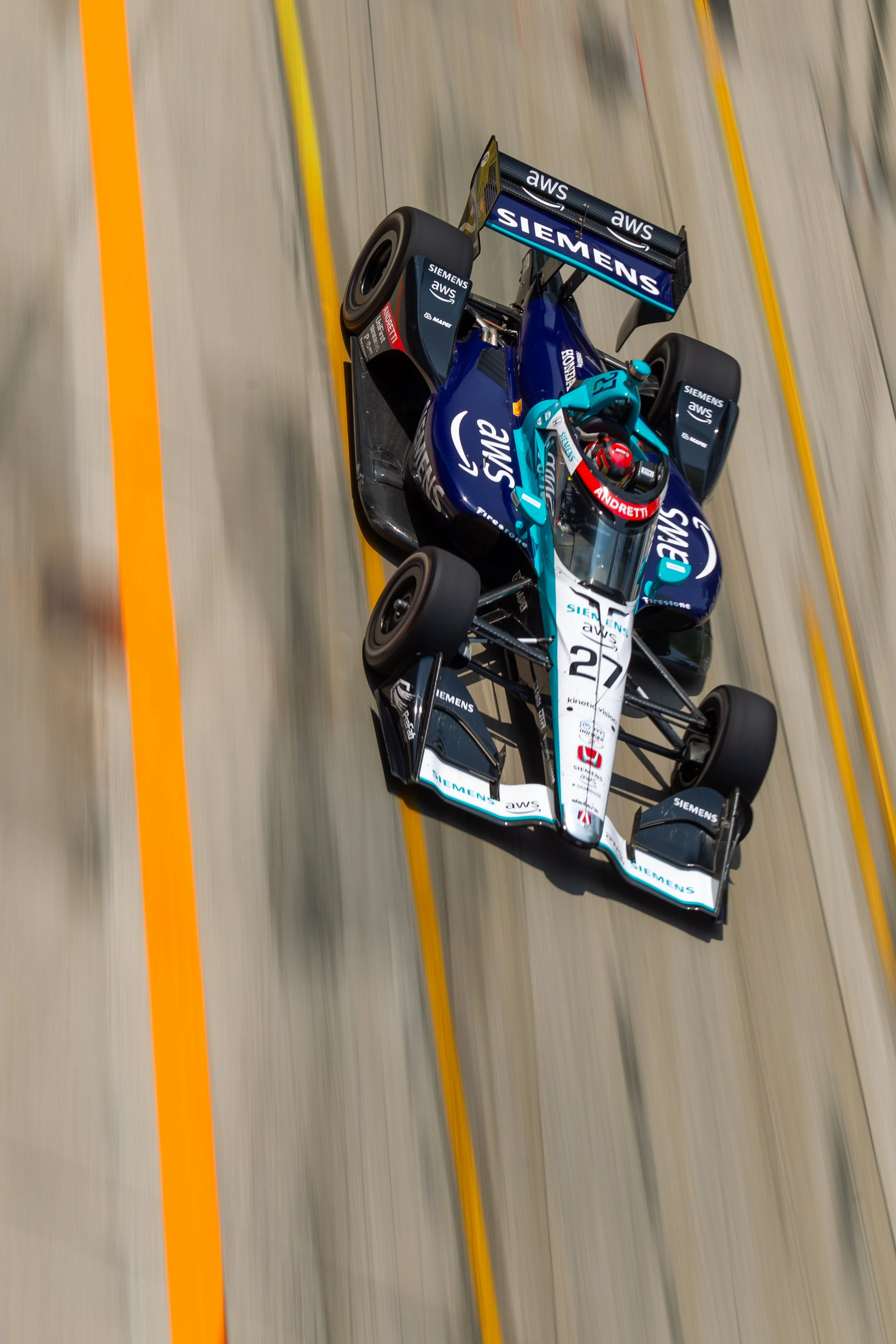 A race car on a track with yellow lines, sponsored by Siemens and AWS, driven by a driver wearing a red helmet.