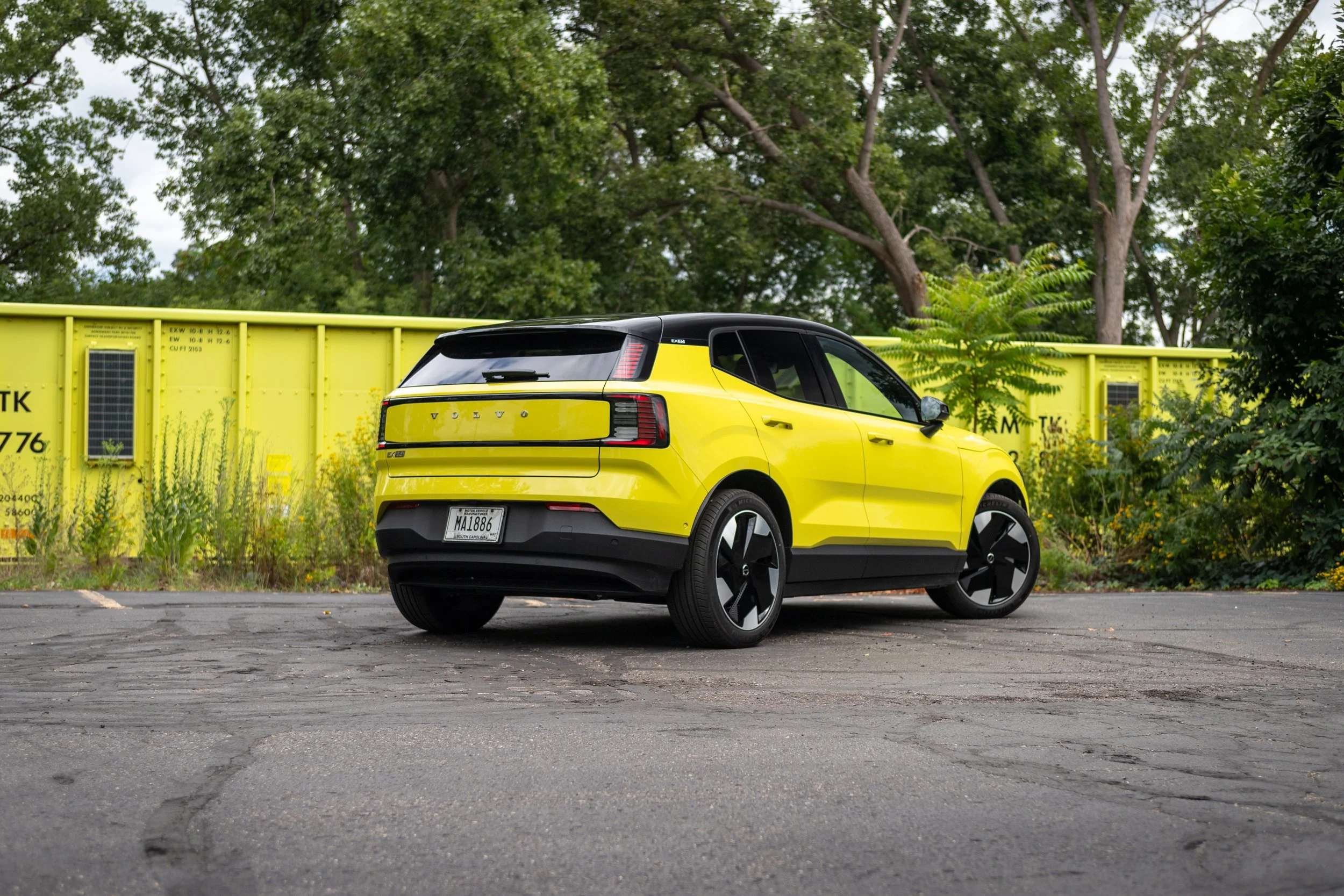 Bright yellow and black electric SUV parked on a cracked asphalt lot outdoors, with lush trees and a yellow construction barrier in the background.