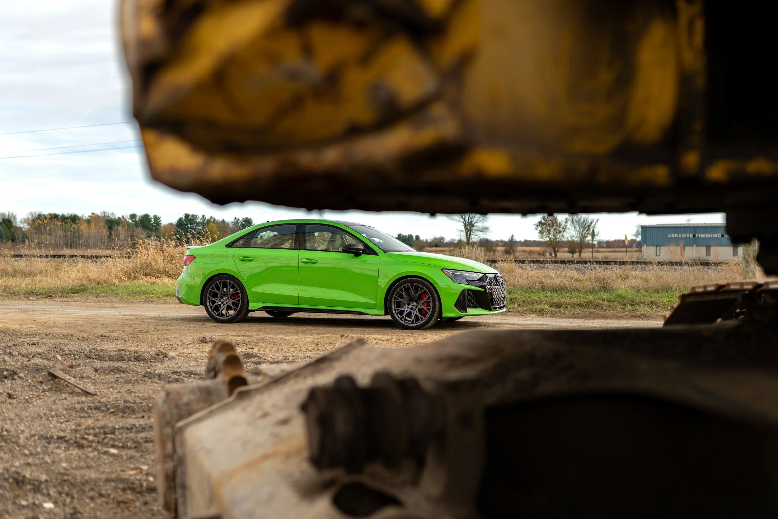 A bright green sports car viewed through a cut-out shape in a pile of logs, with trees and a blue building in the background.