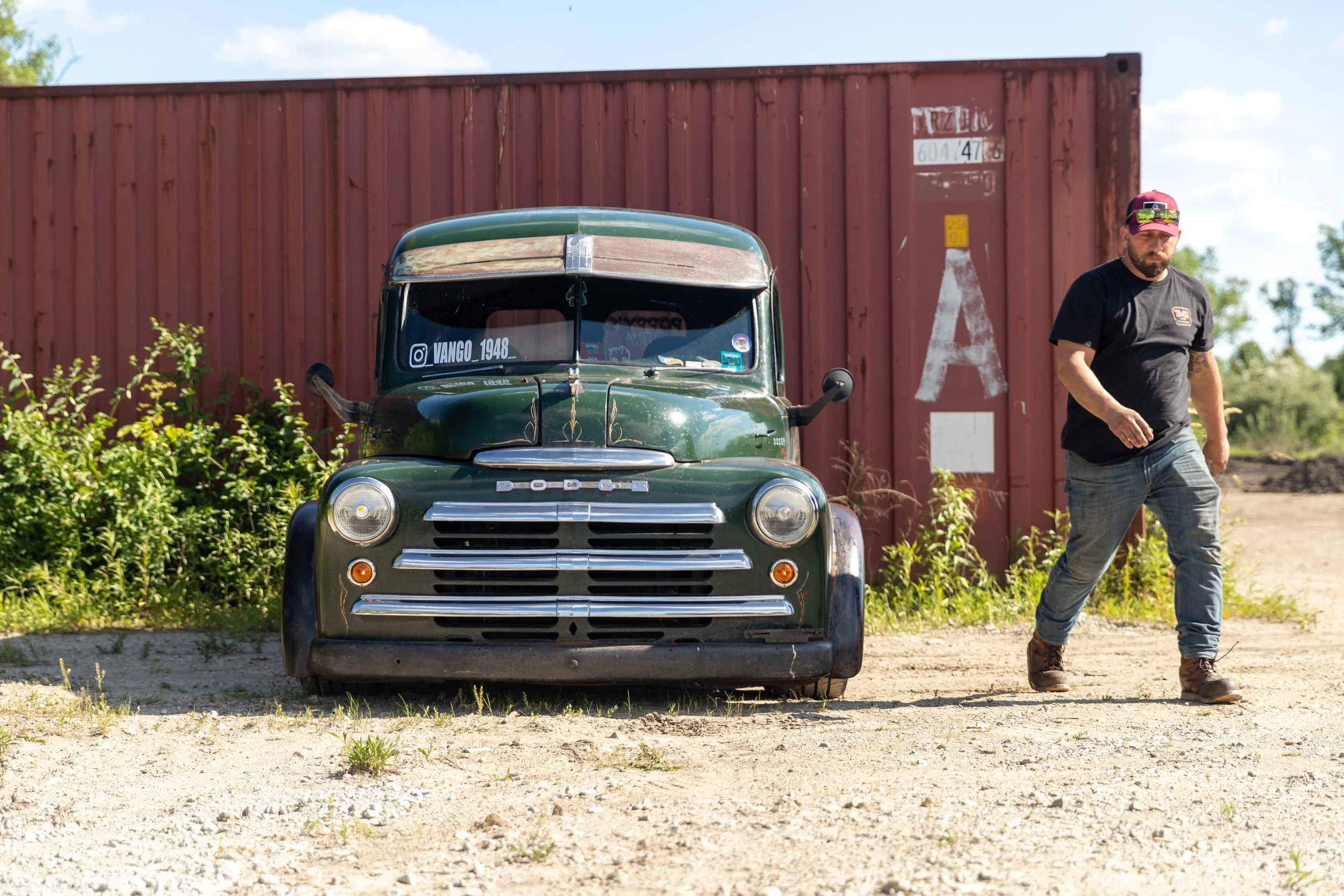 A vintage green Dodge truck parked in front of a rusty shipping container and a man with a beard, wearing a black t-shirt, jeans, brown shoes, and a baseball cap, walking past.