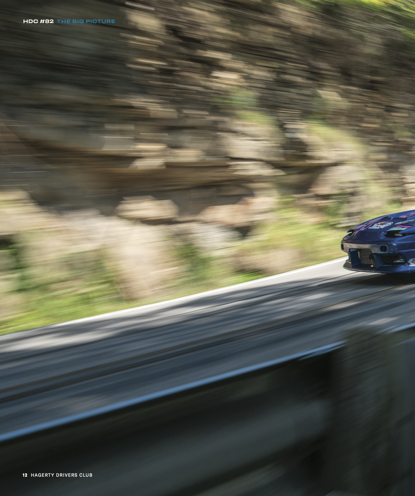 A close-up of a blue race car with sponsor decals speeding along a highway, with a blurred background of rocks and greenery.
