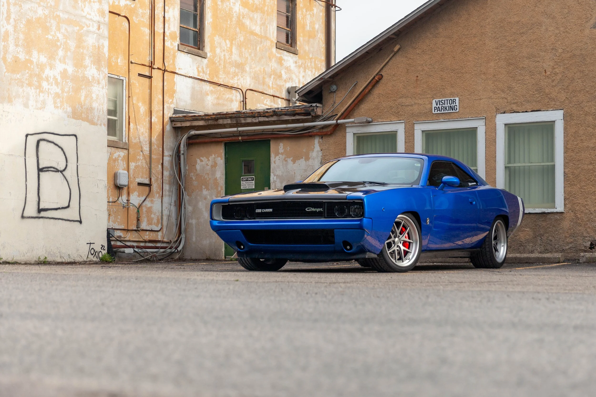 A blue Dodge Challenger car parked in front of a worn, yellow and beige building with graffiti and pipes.