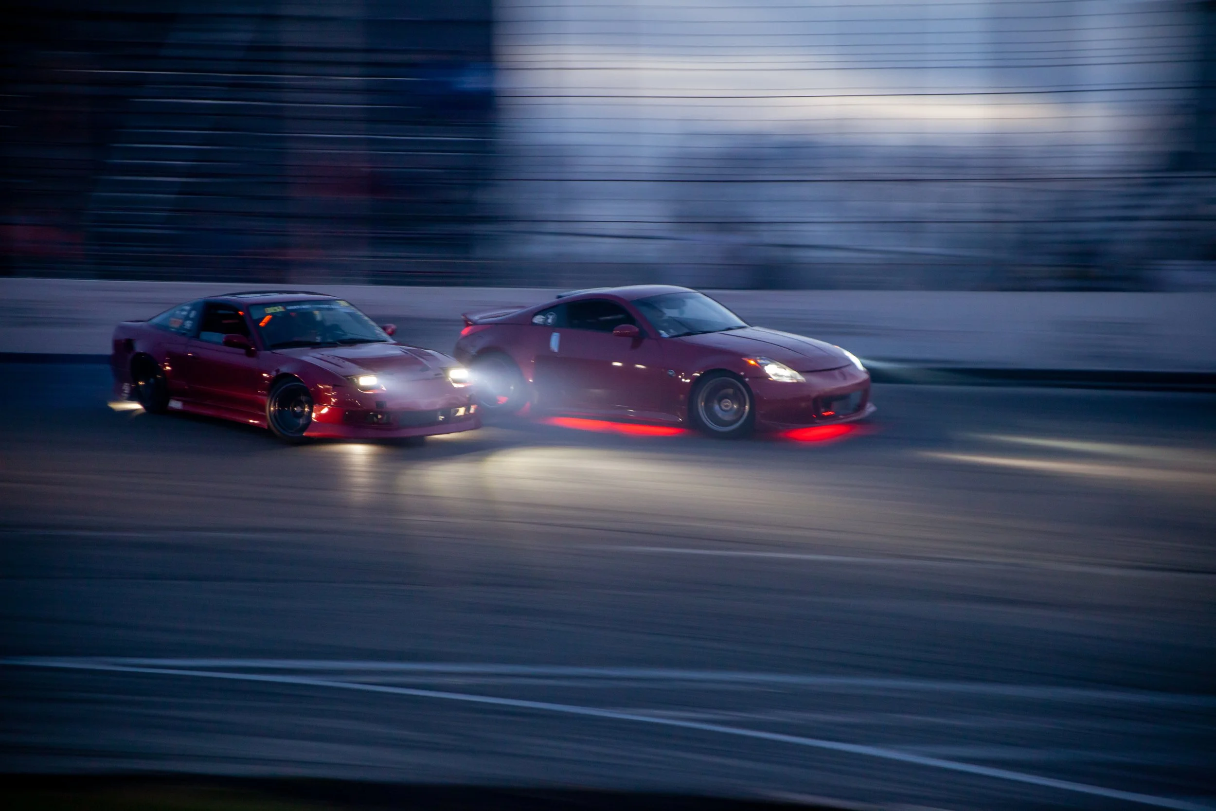 Two red sports cars racing side by side on a track at dusk, with a blurred background indicating high speed.