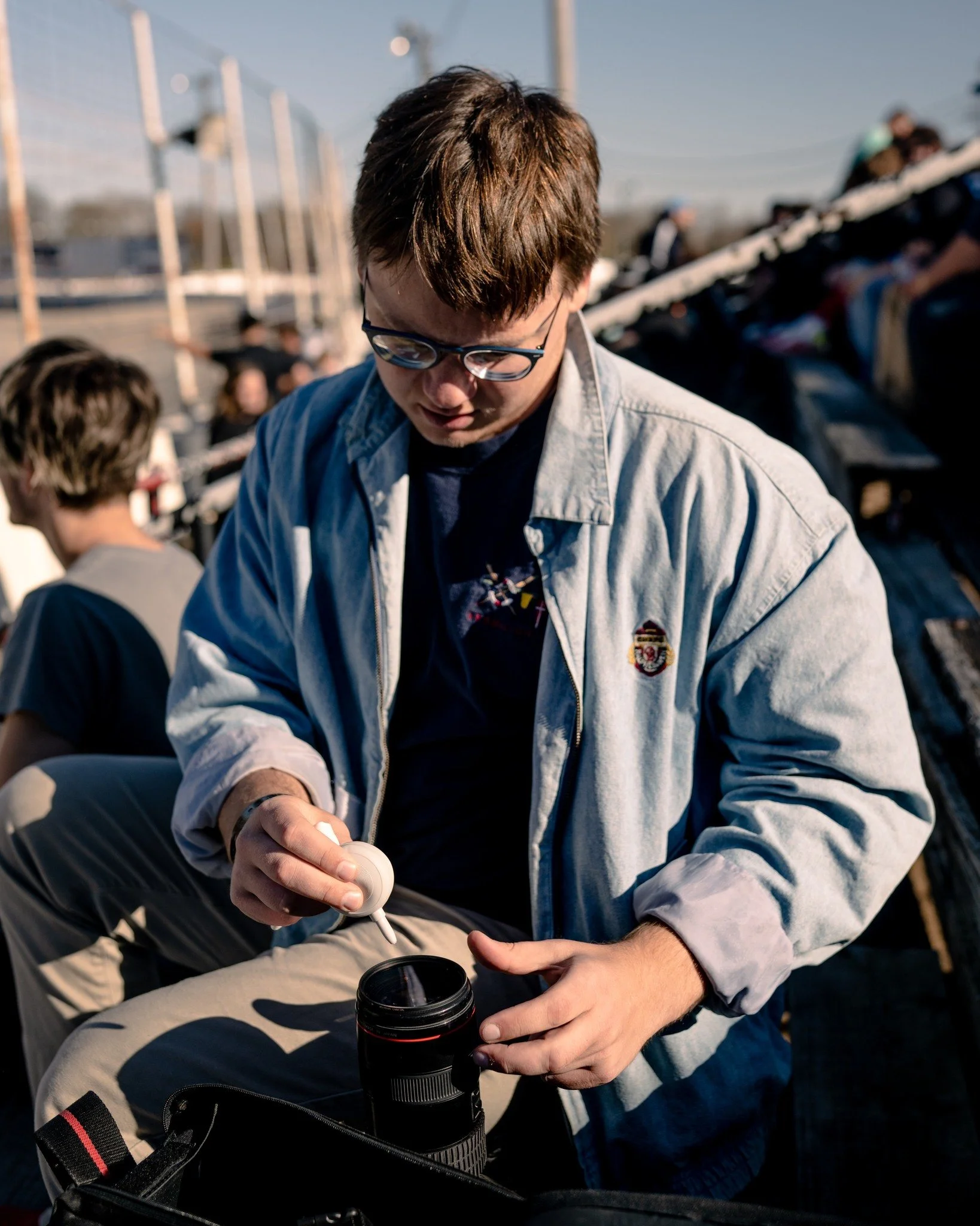 A young man with glasses wearing a light-colored jacket, sitting outdoors, opening a black thermos, with a lens in front of him and a backpack nearby, with a crowd of people in the background under a clear blue sky.