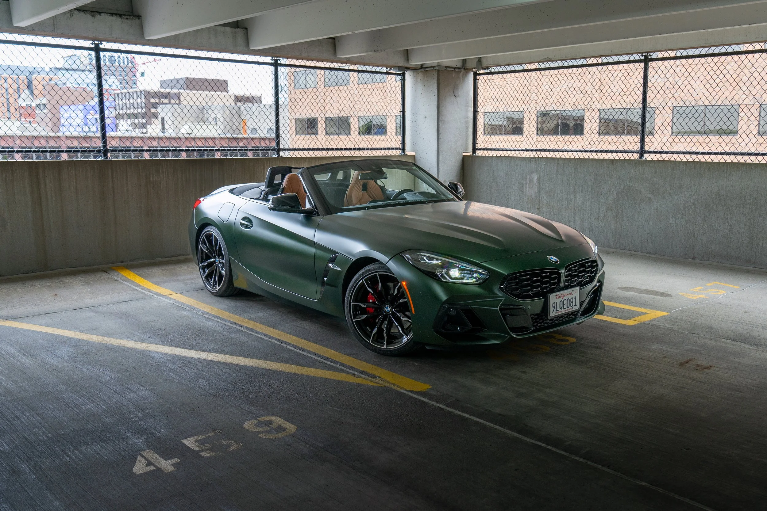 A dark green BMW convertible car parked in an indoor parking garage with metal railings and city buildings in the background.
