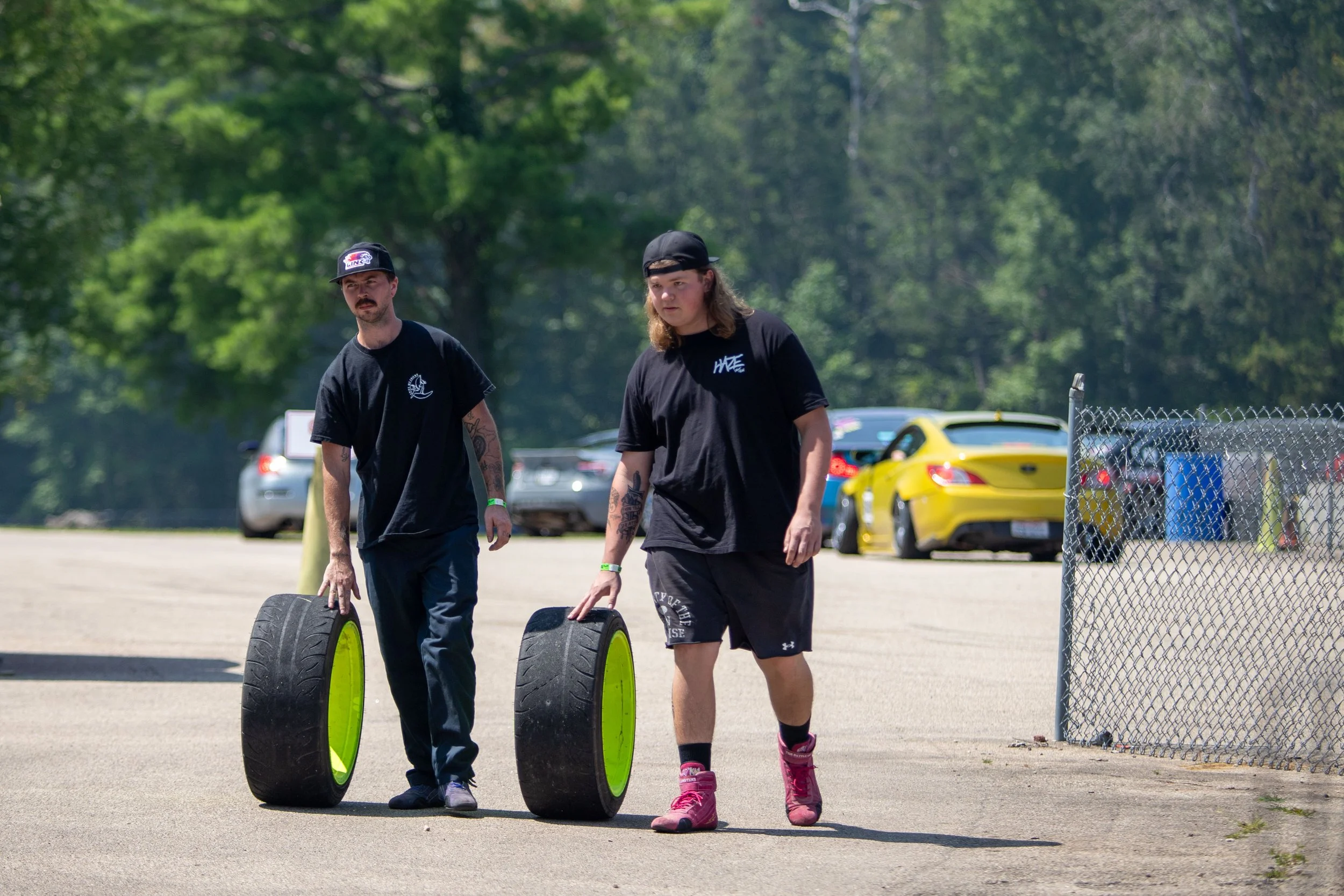Two young men are walking outdoors, each holding a large tire with yellow rims. One is wearing a black short-sleeve shirt and a black cap, and the other is dressed in black shorts, a black T-shirt, and a backward black cap. They are on a paved surfac