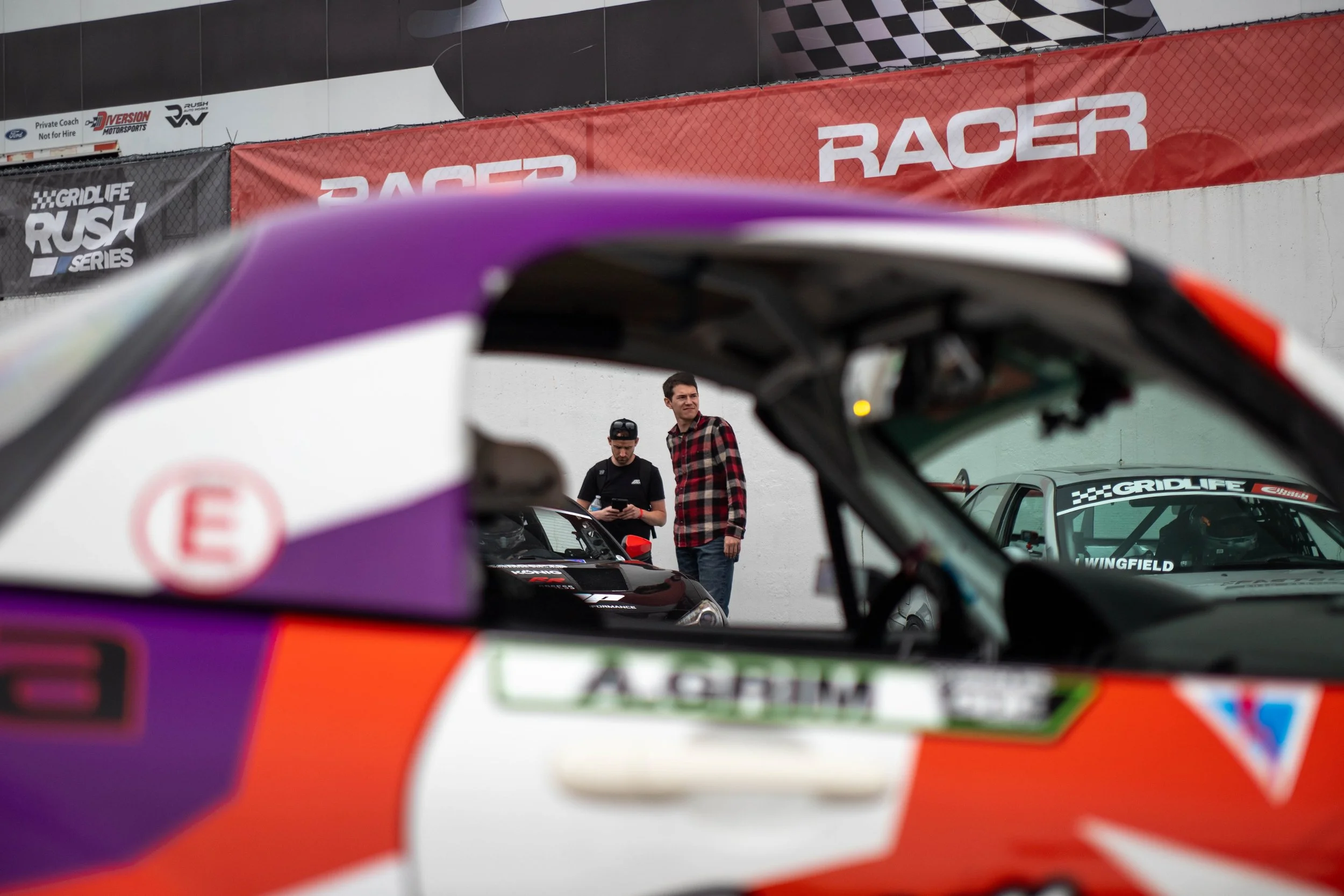 View of race cars and two people standing near the cars at a racing event, seen through the windshield of a colorful race car.
