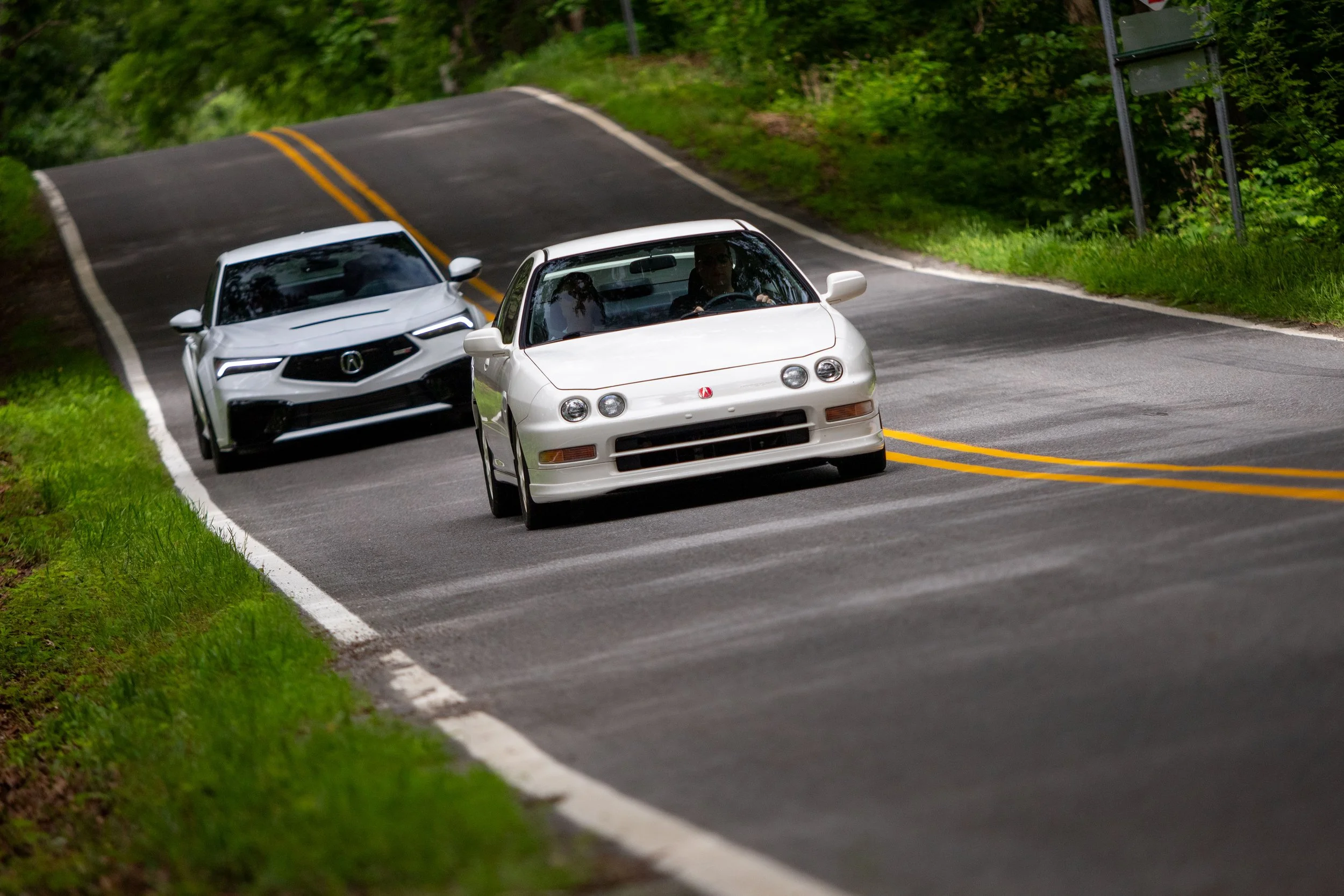 Two white cars driving down a winding, two-lane road surrounded by green trees and grass.