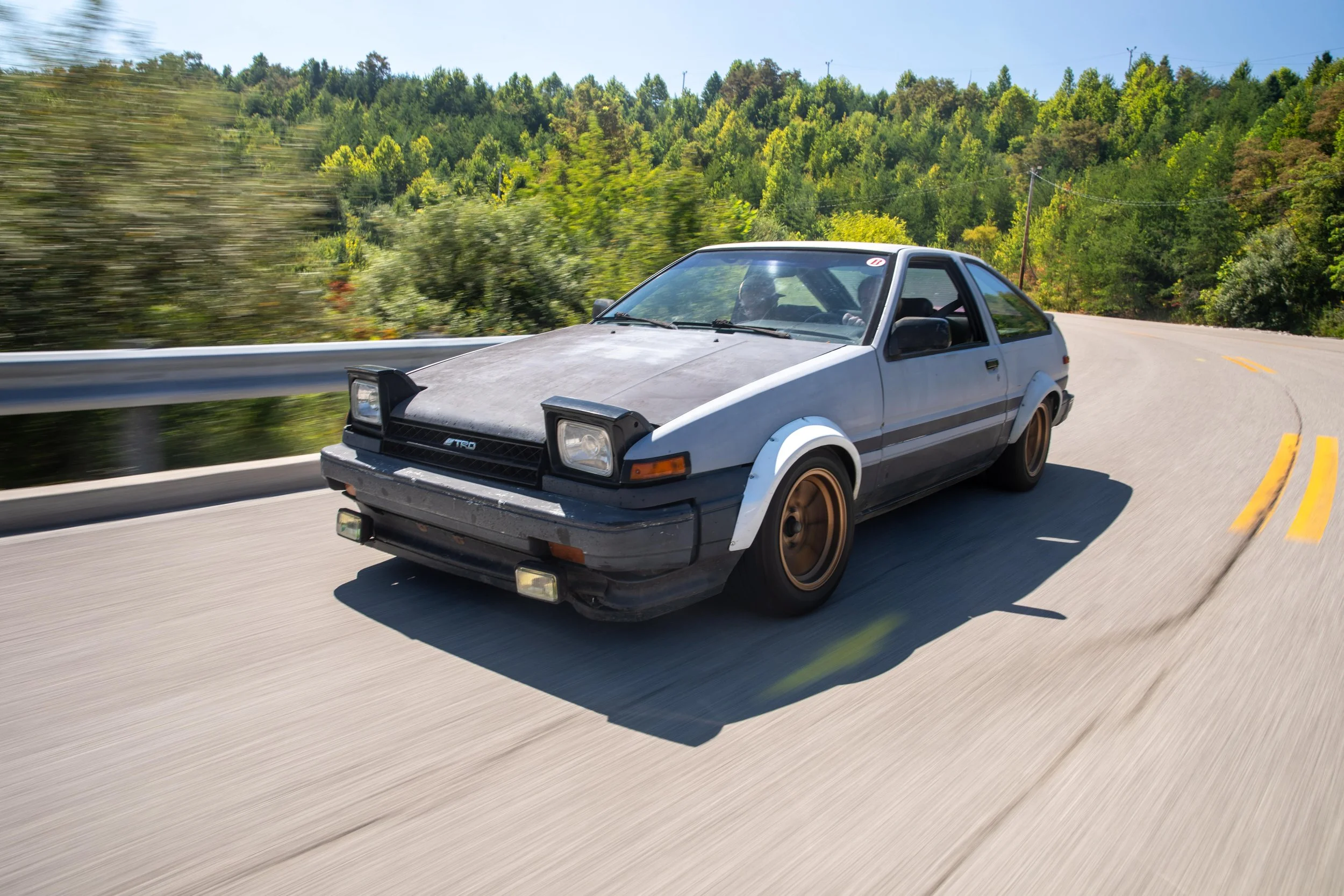 A silver vintage hatchback car with a black hood is driving on a winding road surrounded by lush green trees and hills.