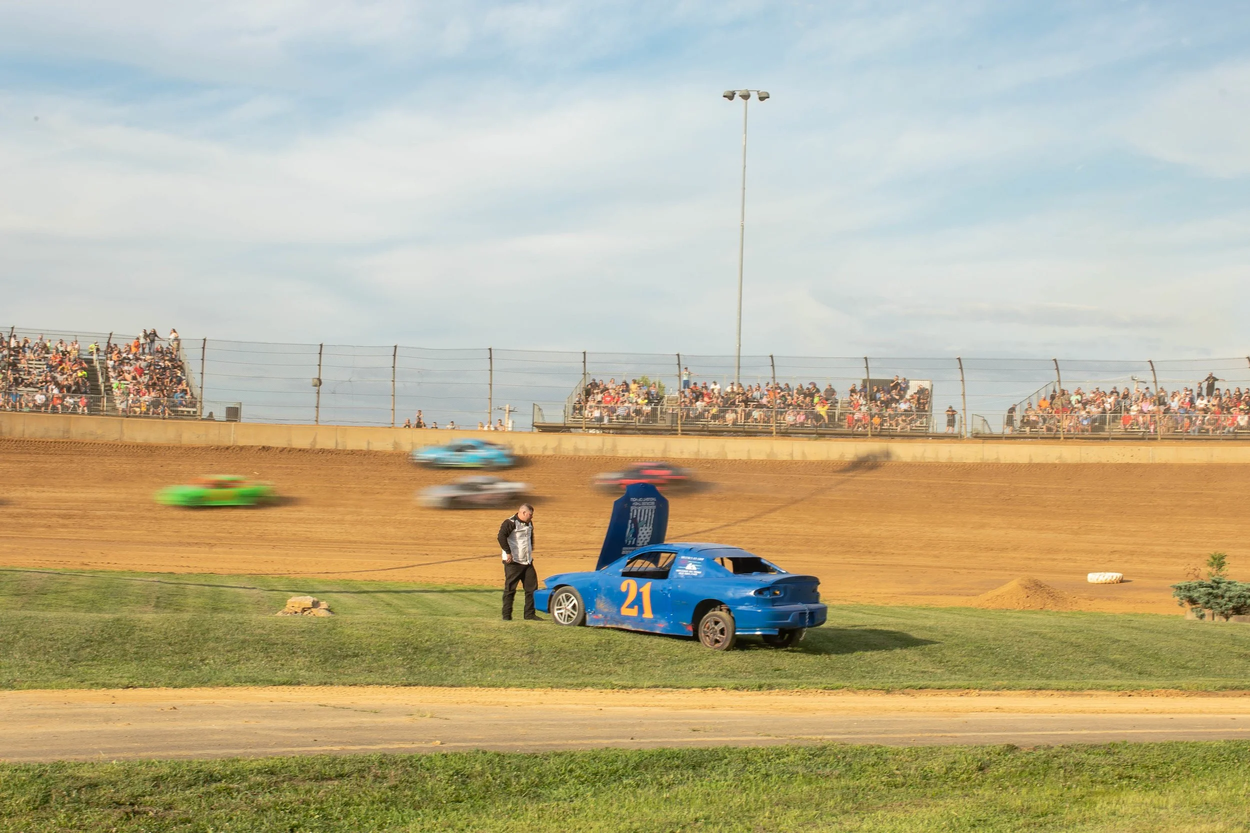 Race car number 21 on an asphalt track, with blurred racing cars in the background on a dirt track, and spectators watching from bleachers behind a fence.