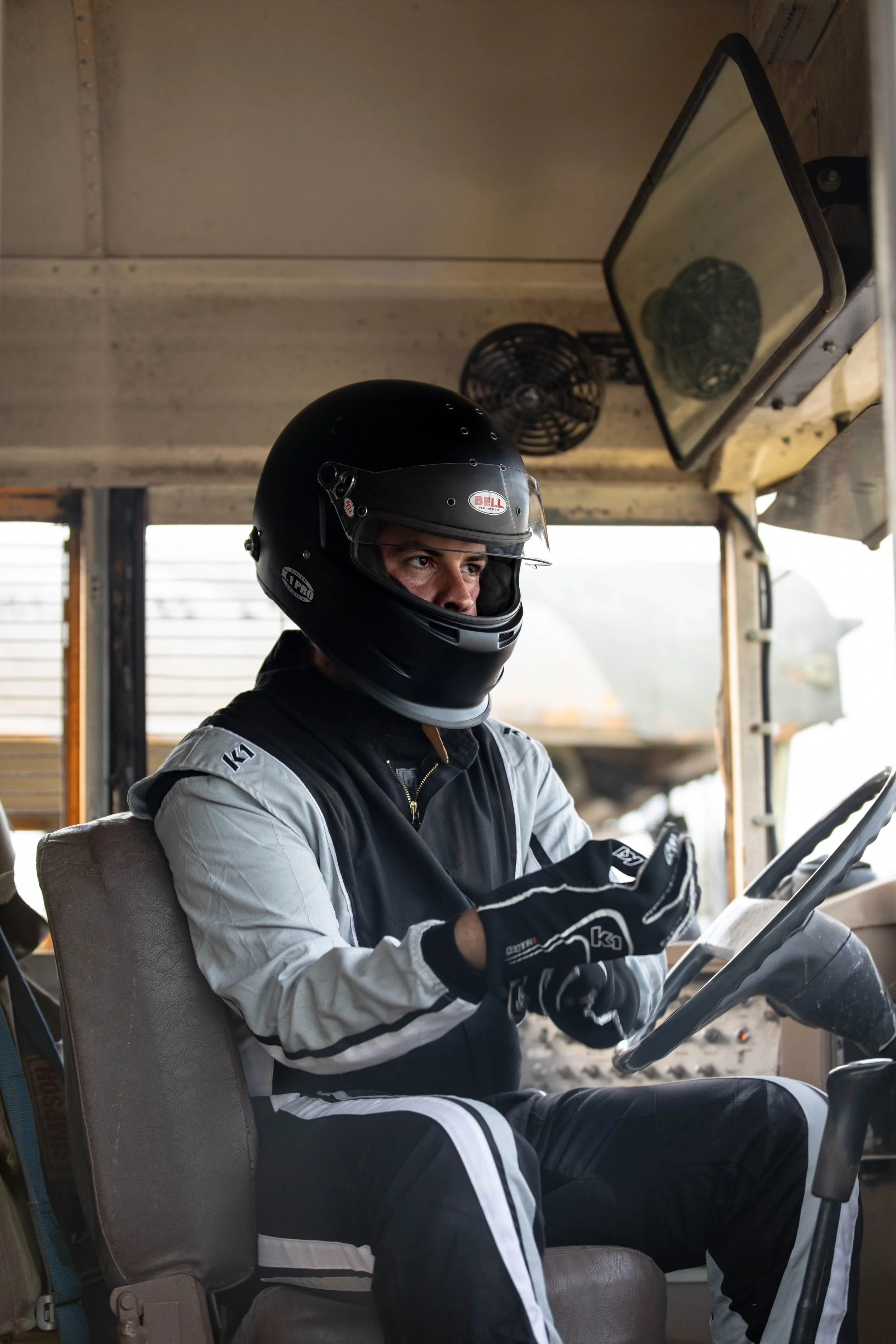 A person wearing a black full-face helmet with a clear visor, seated in an electric golf cart. The individual is dressed in a white and black racing suit, with gloves and black pants with white stripes, and appears to be looking at a device in their 