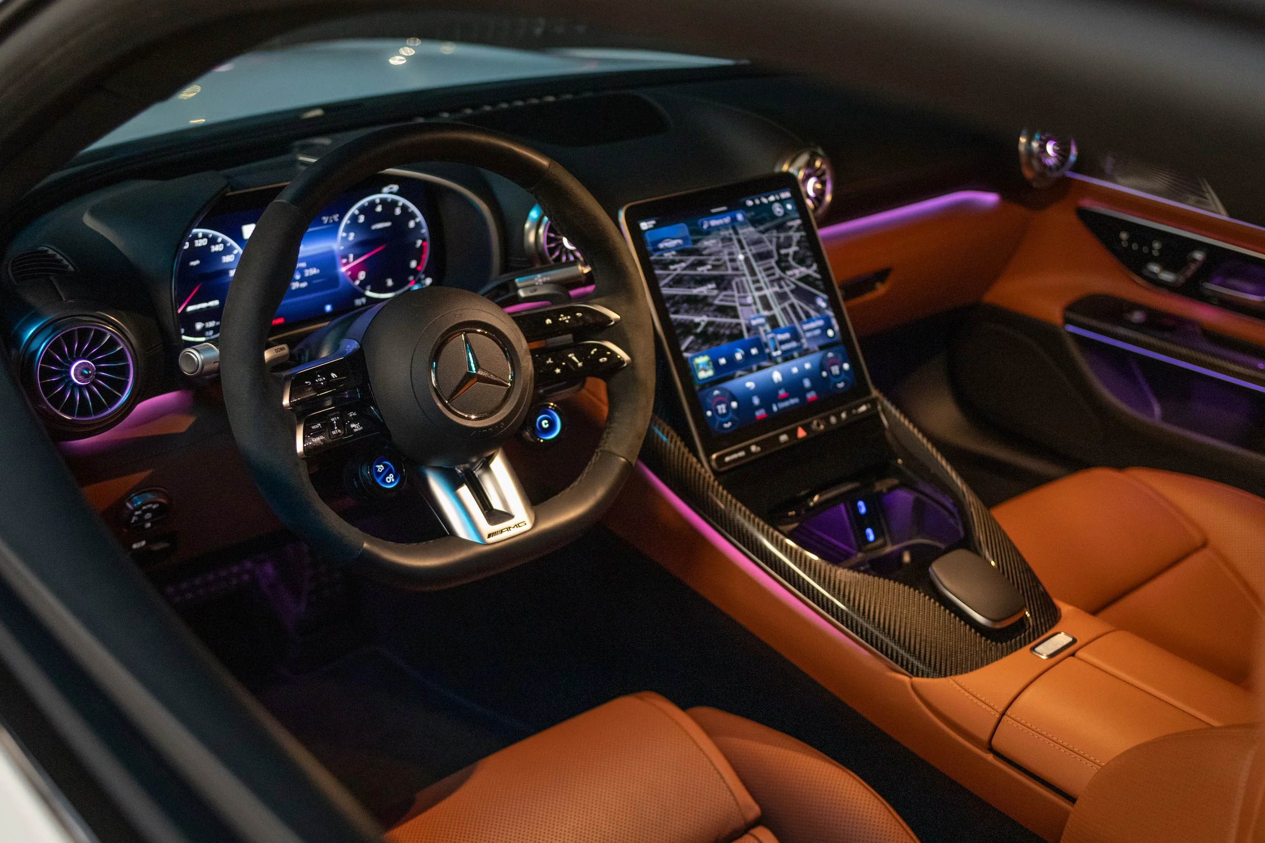 Interior of a luxury Mercedes-Benz vehicle with a digital dashboard, a large touchscreen display showing a navigation map, illuminated ambient lighting, and tan leather seats.