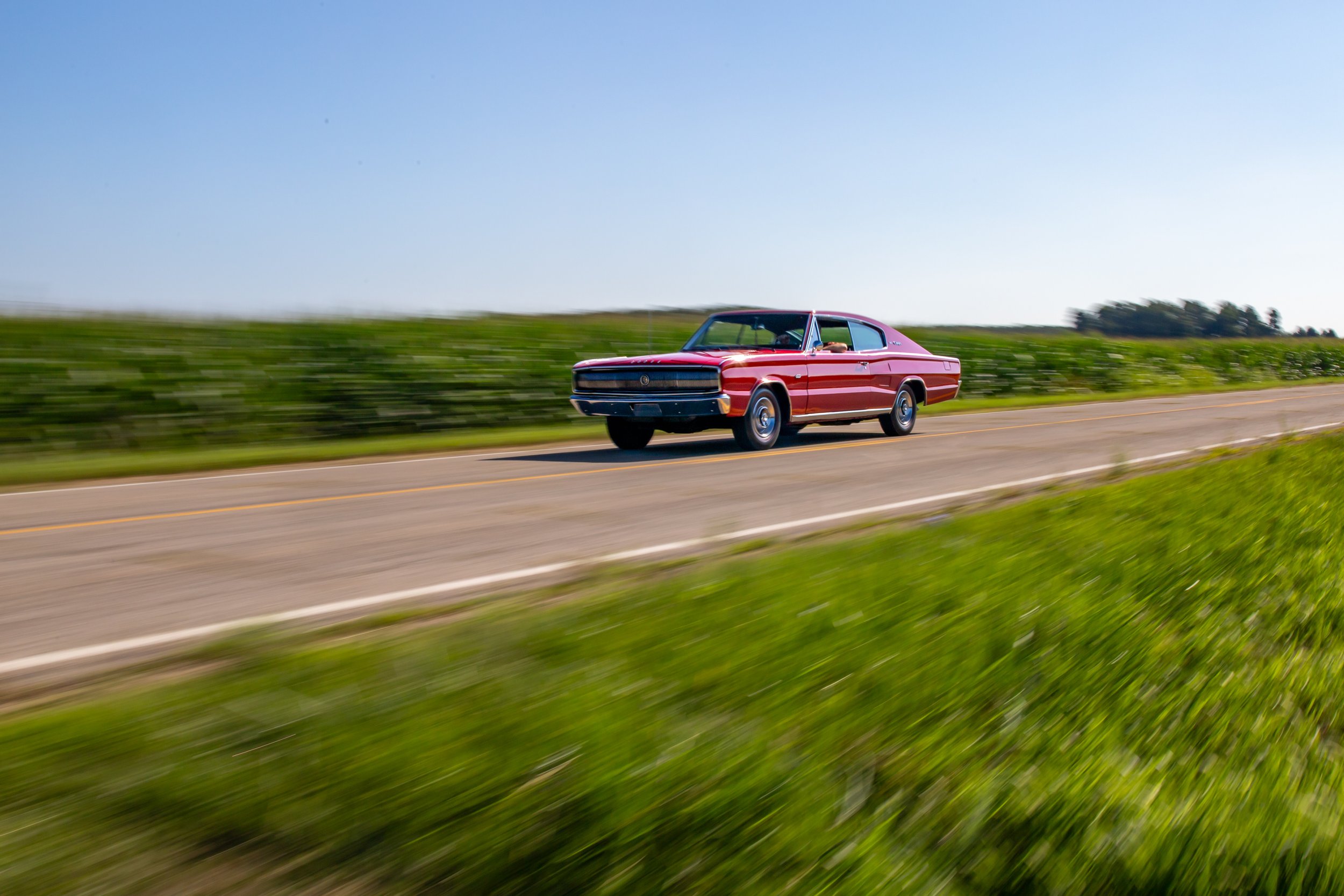 A red vintage car driving on a rural highway through green fields with a clear blue sky in the background.