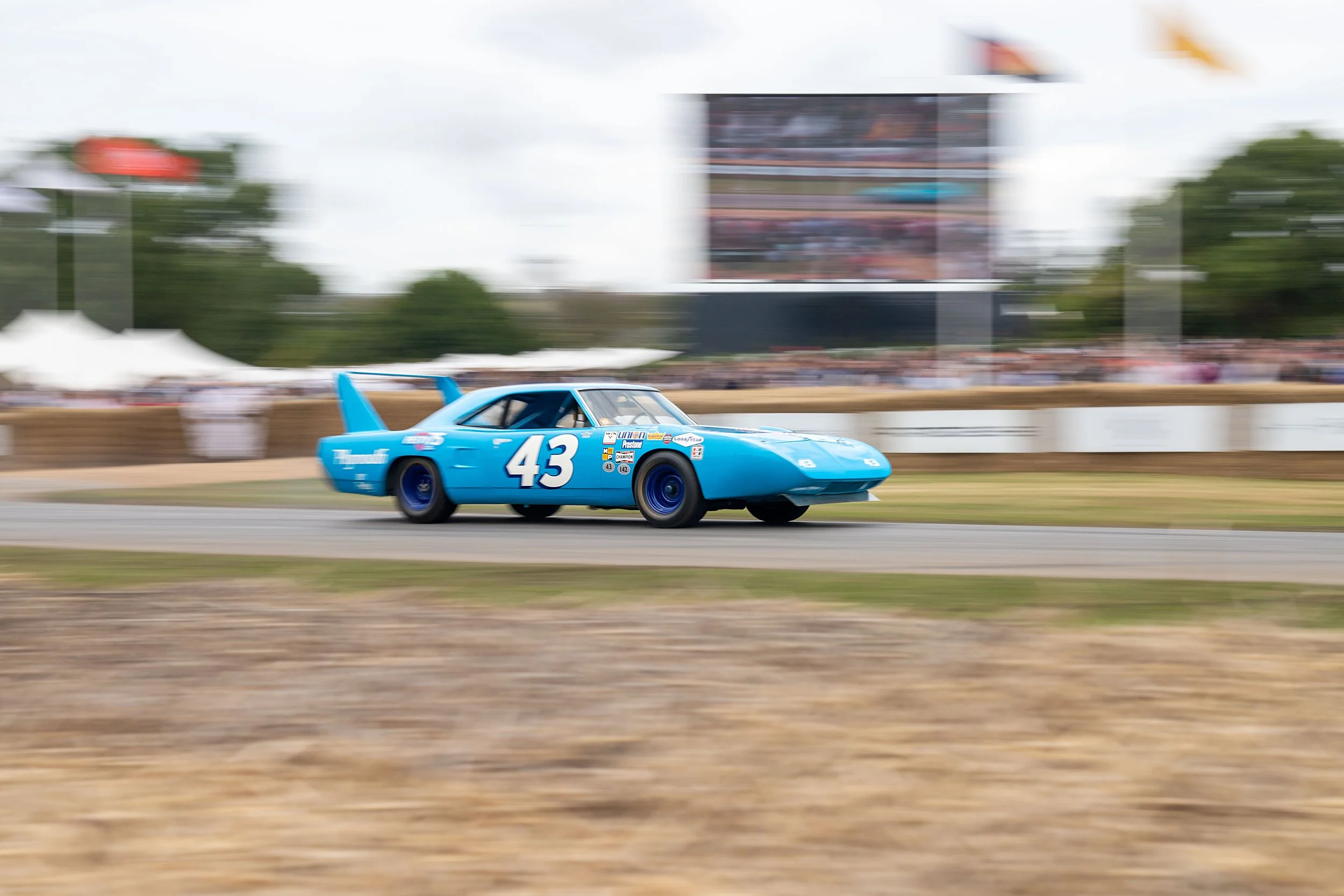 A blue race car with the number 43 on its side speeding on a racetrack, with a digital scoreboard and trees in the background, captured with motion blur.