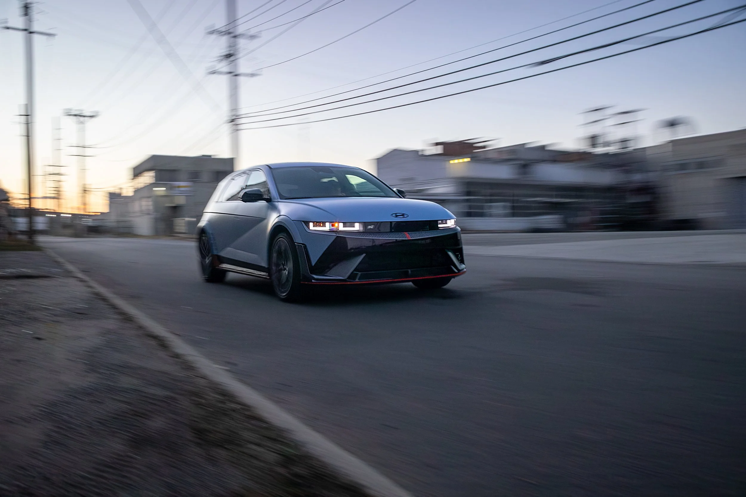 A sleek gray sports car driving on an urban street at sunset, with motion blur in the background and power lines overhead.
